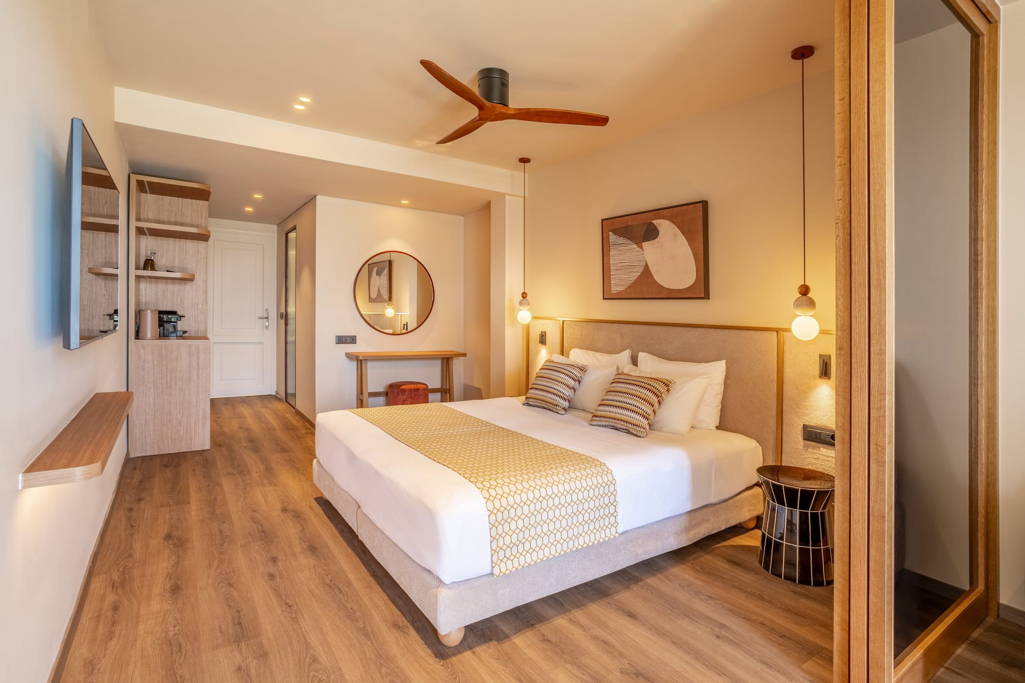 Modern hotel bedroom with king bed, wooden floor, beige walls, and ceiling fan.