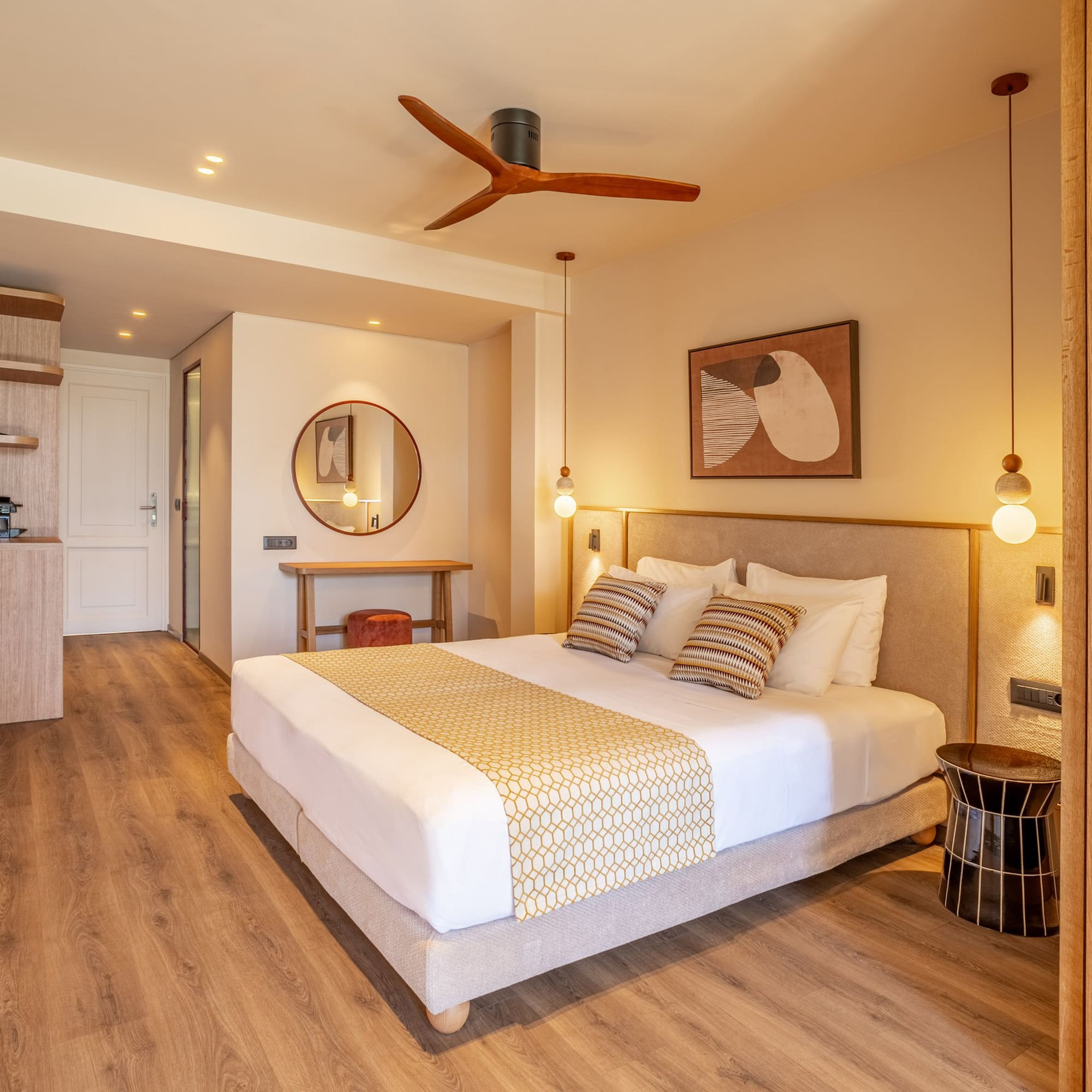 Modern hotel bedroom with king bed, wooden floor, beige walls, and ceiling fan.