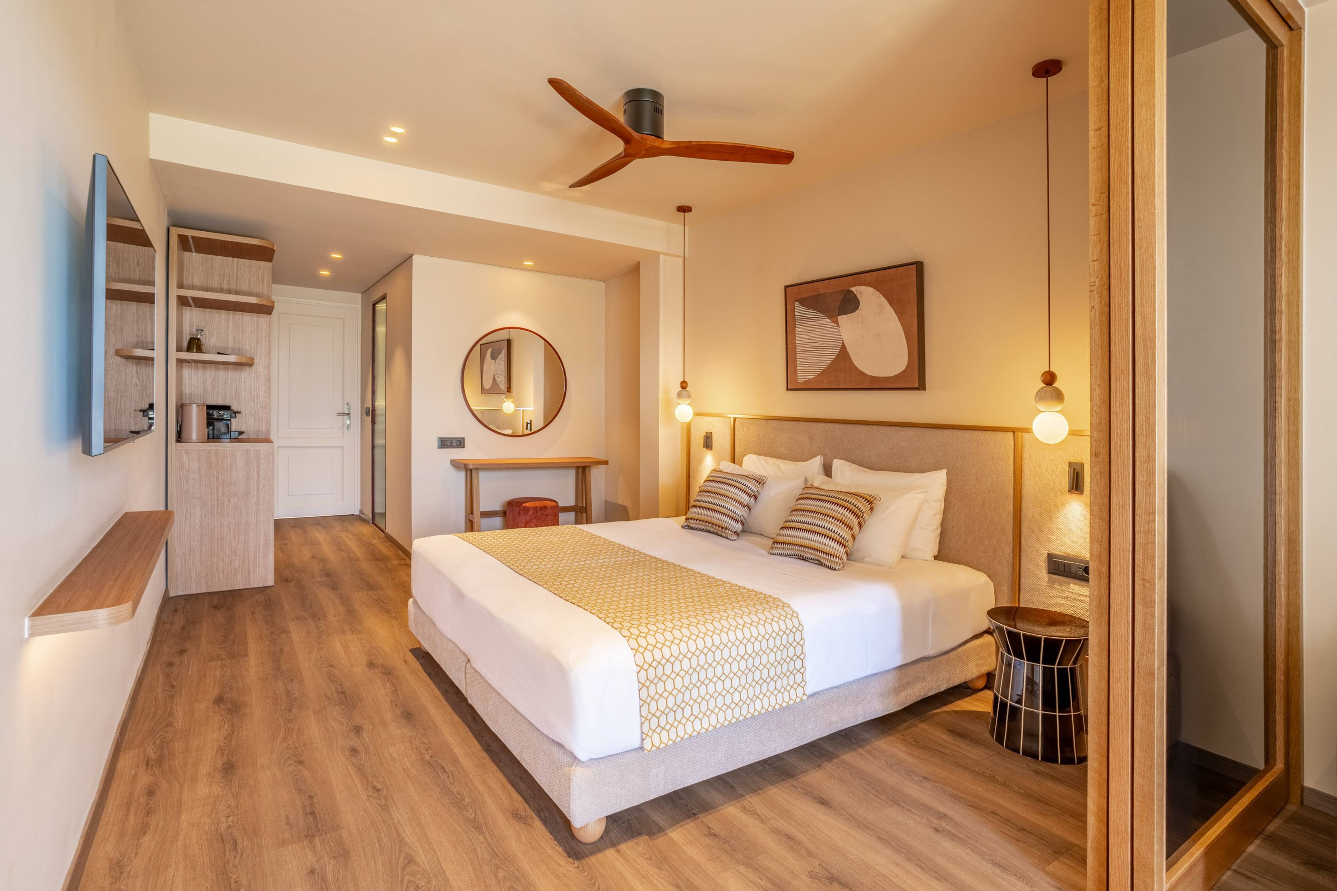 Modern hotel bedroom with king bed, wooden floor, beige walls, and ceiling fan.