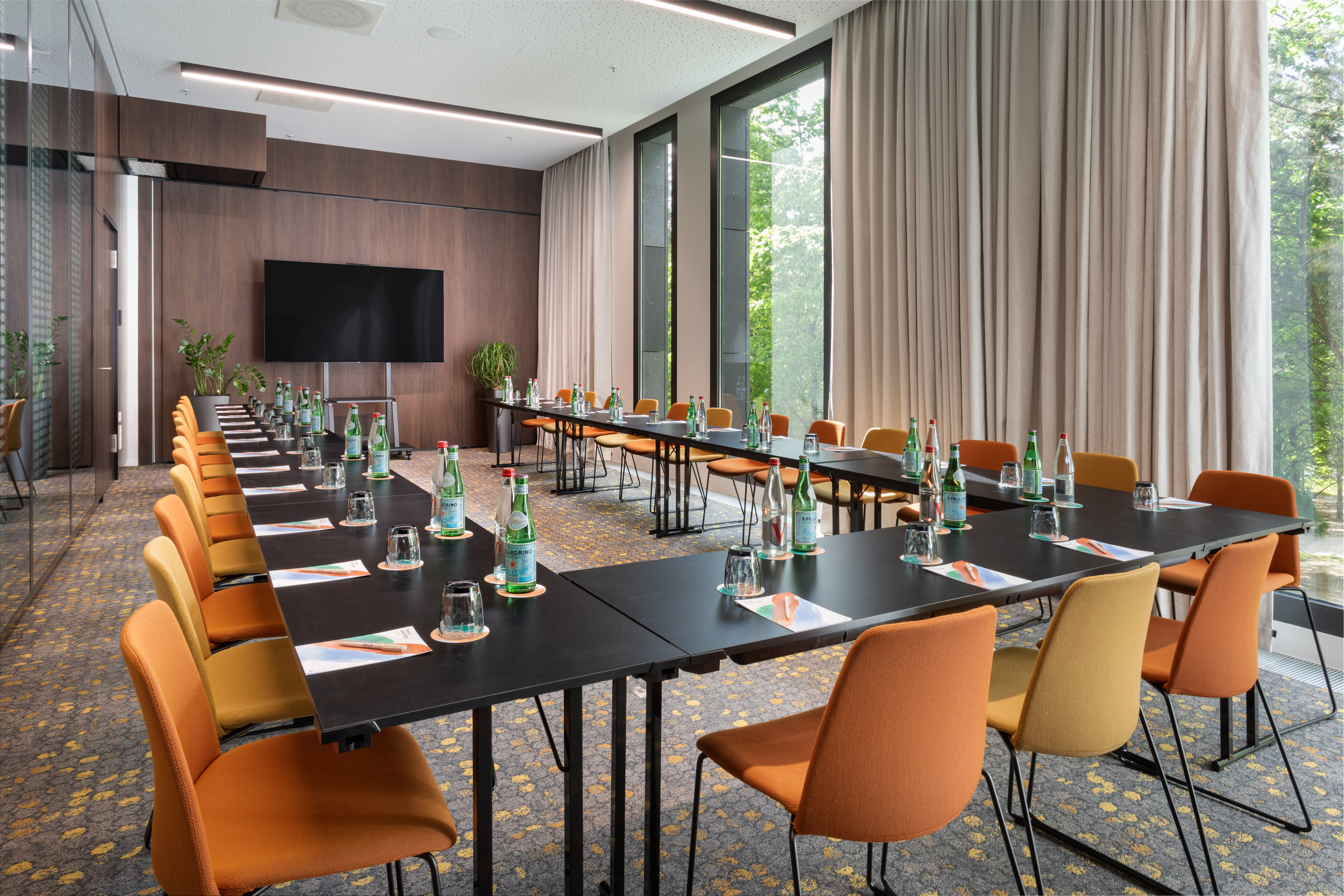 Modern U-shaped meeting room, orange chairs, large TV, and sunlit windows.