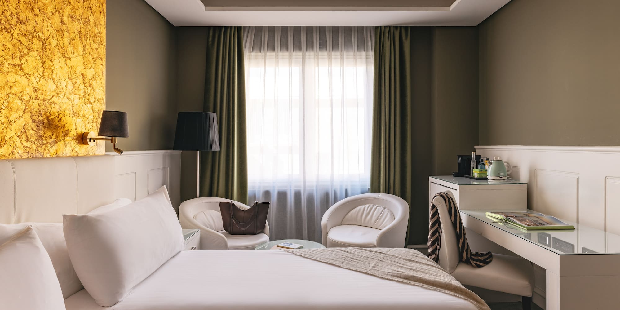 Suite de hotel de lujo: cama, escritorio, sillones blancos y pared dorada.