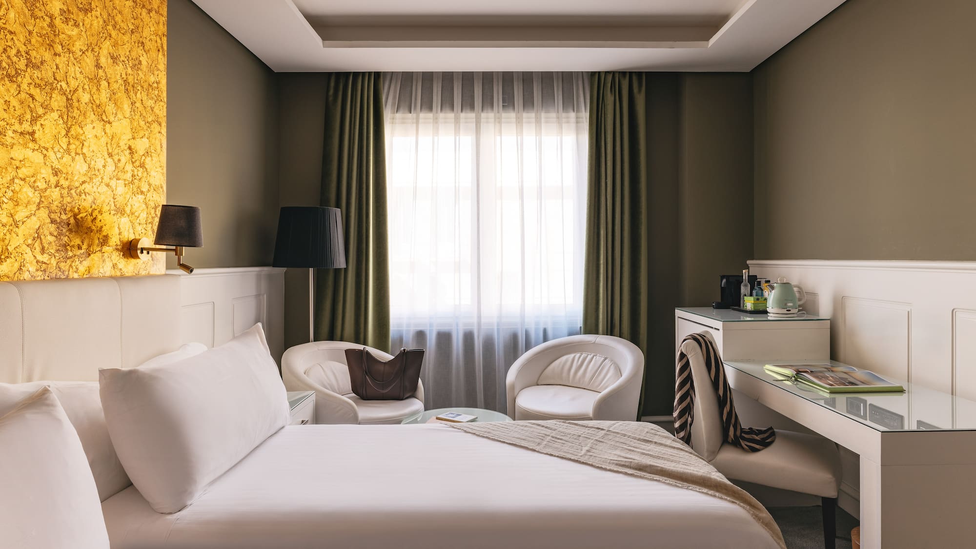Suite de hotel de lujo: cama, escritorio, sillones blancos y pared dorada.