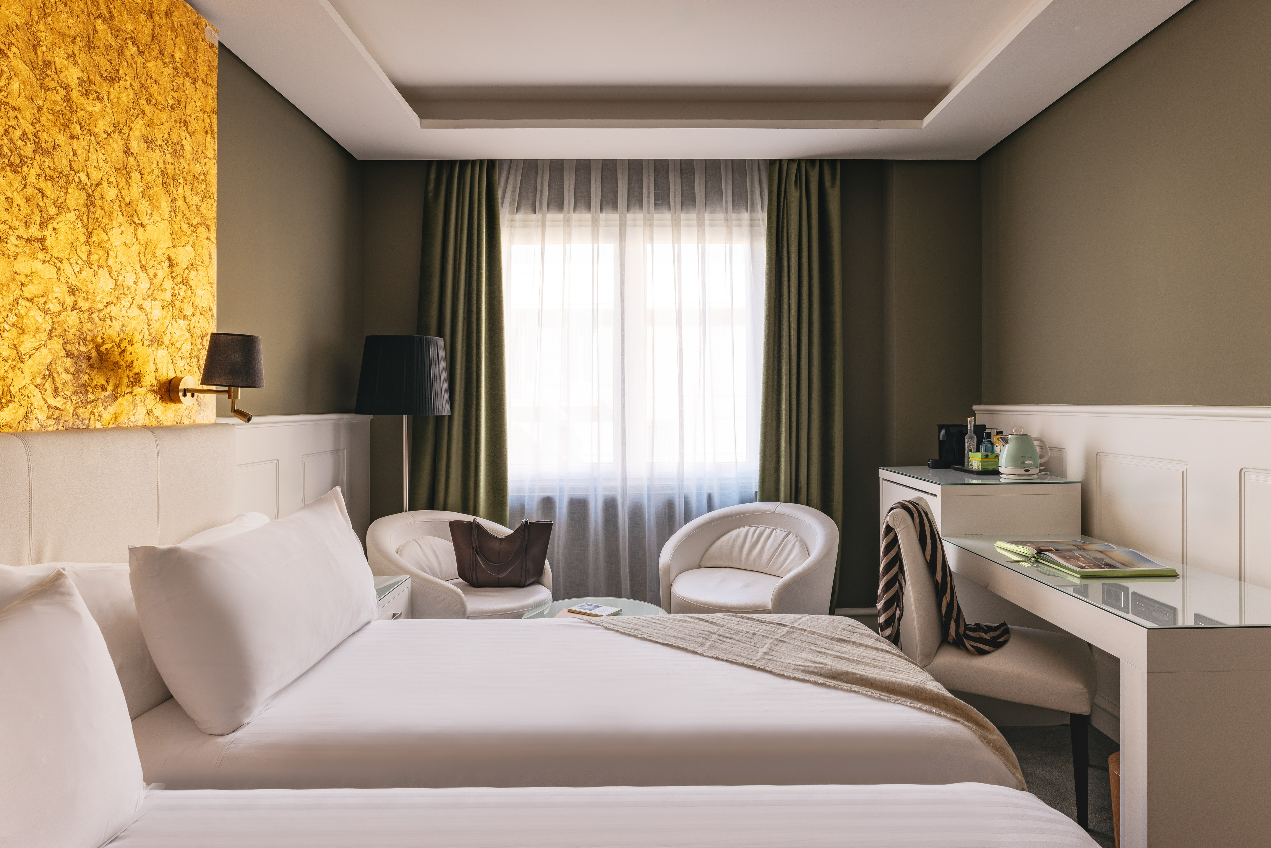 Suite de hotel de lujo: cama, escritorio, sillones blancos y pared dorada.
