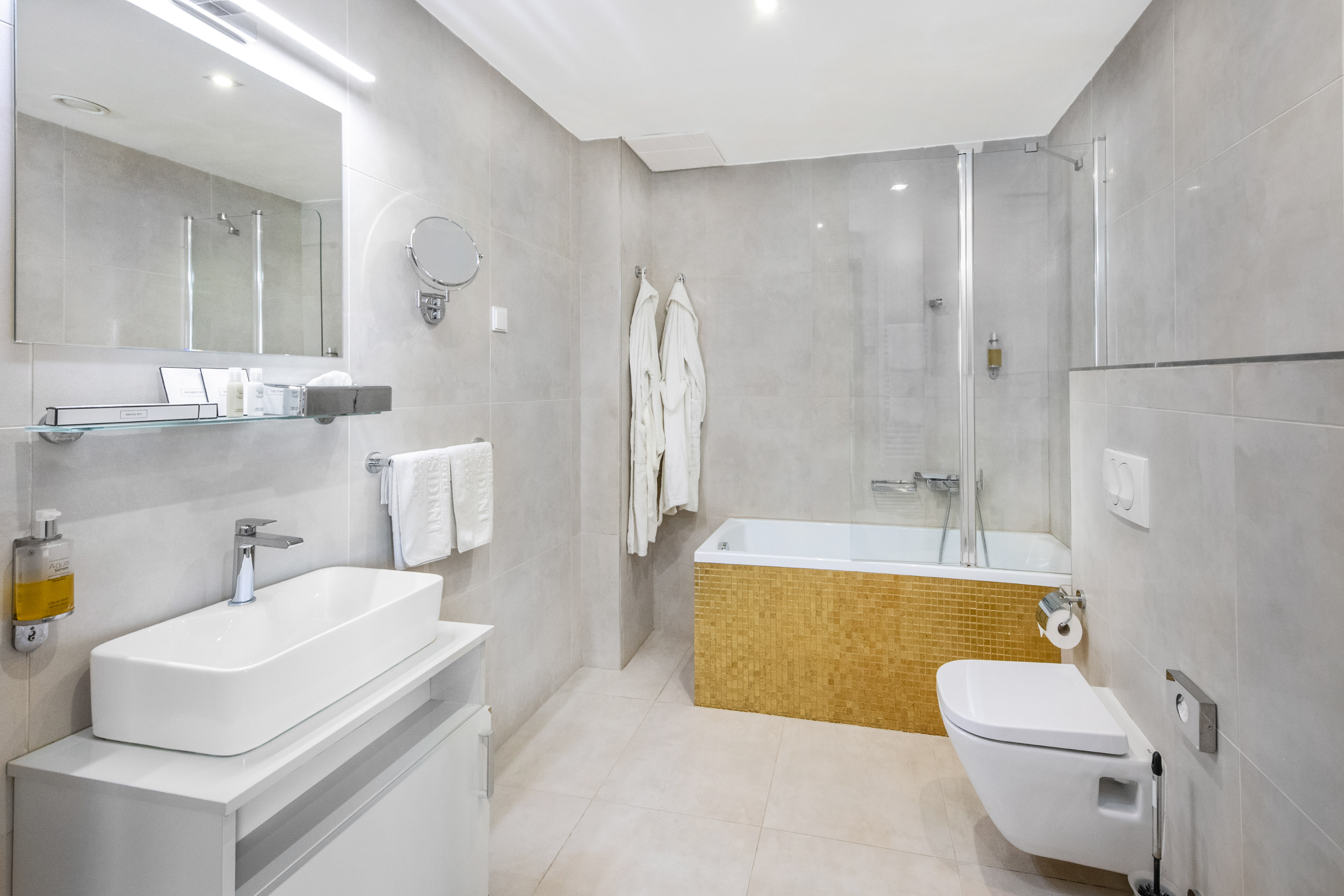 Modern bathroom with white vanity, gold mosaic tub, glass shower, and toilet.