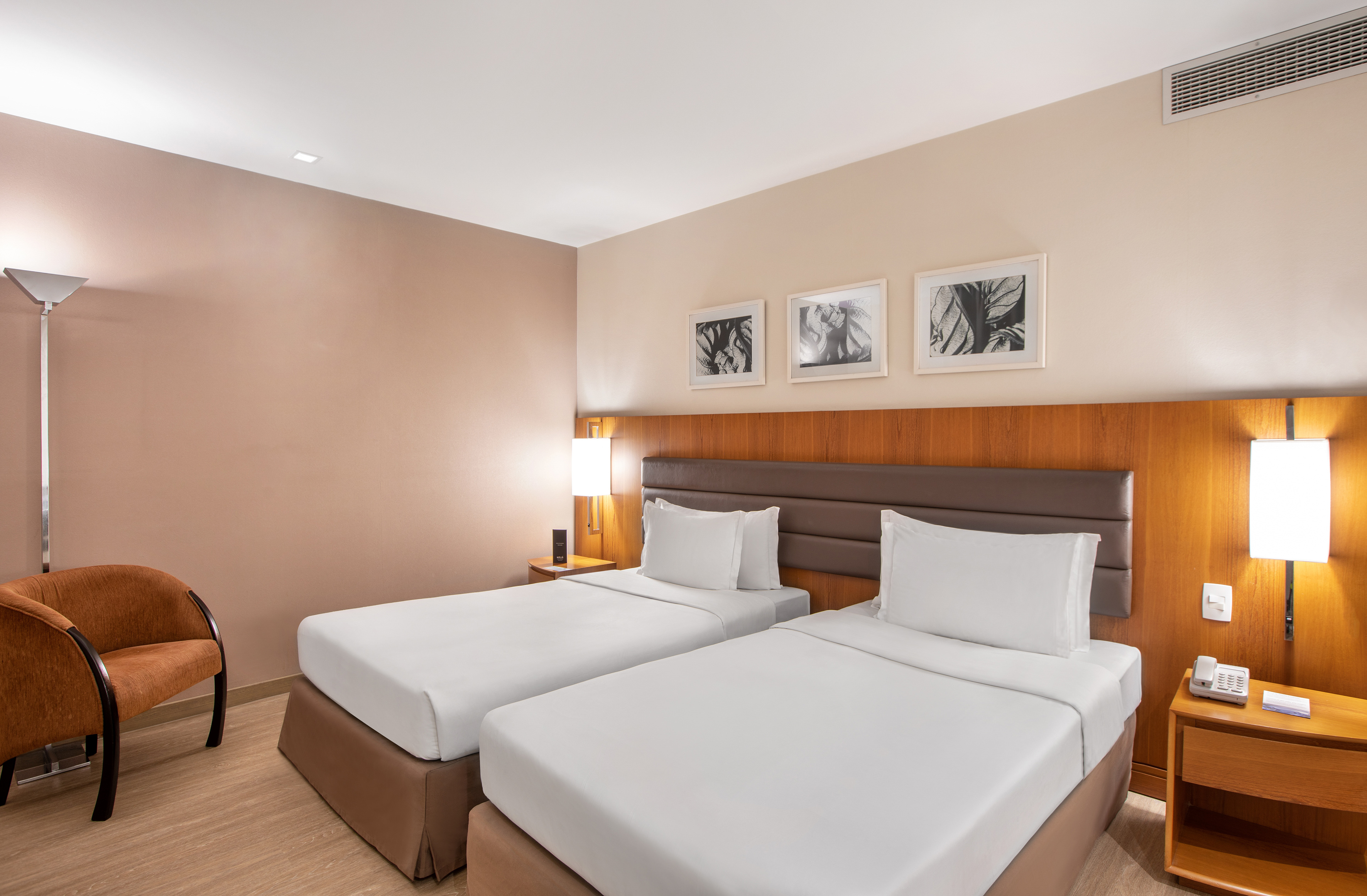 Hotel guest room with two single beds, wooden headboard, and an accent chair.