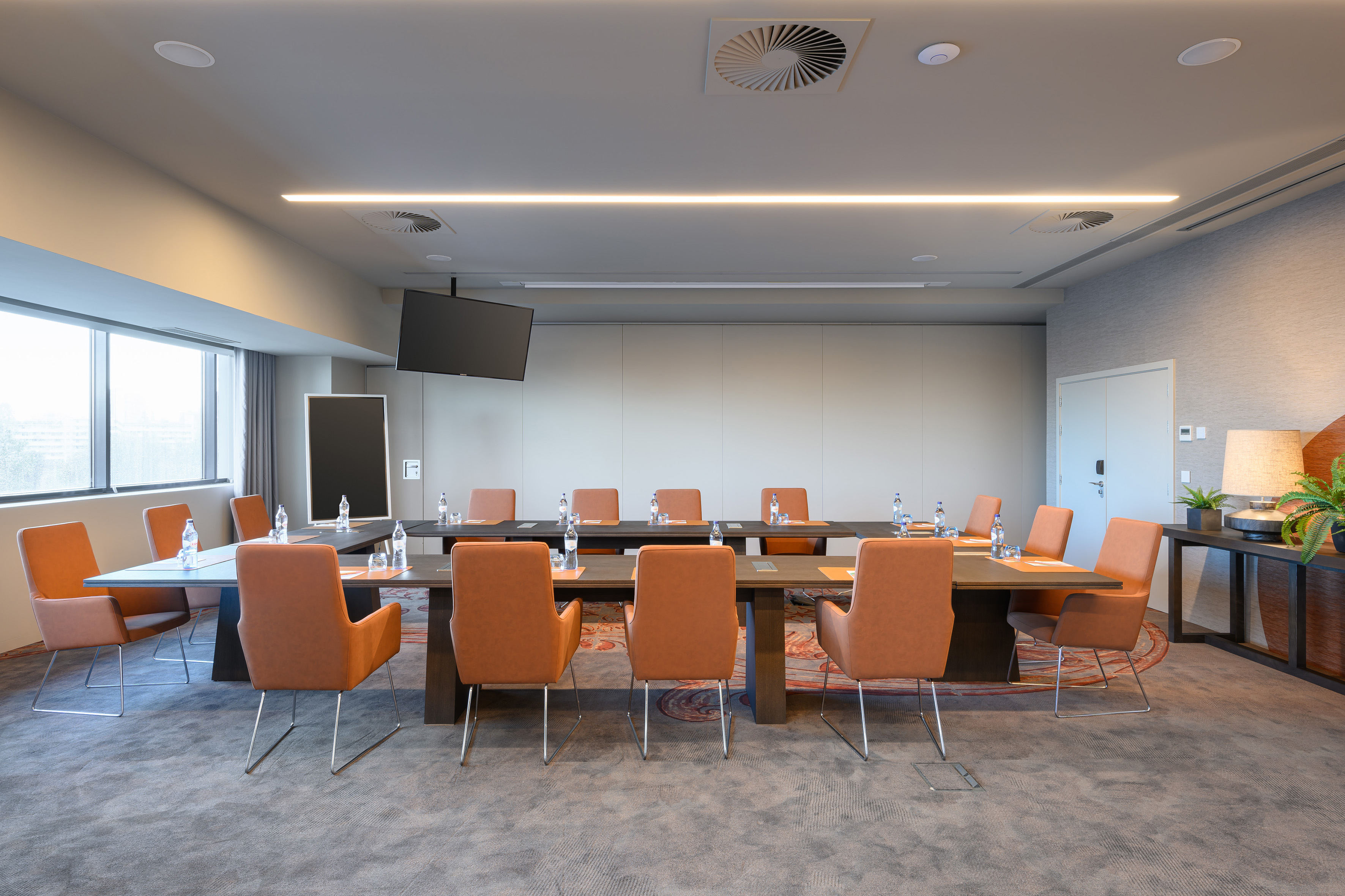 Modern conference room with U-shaped table, orange chairs, large window, and TV.