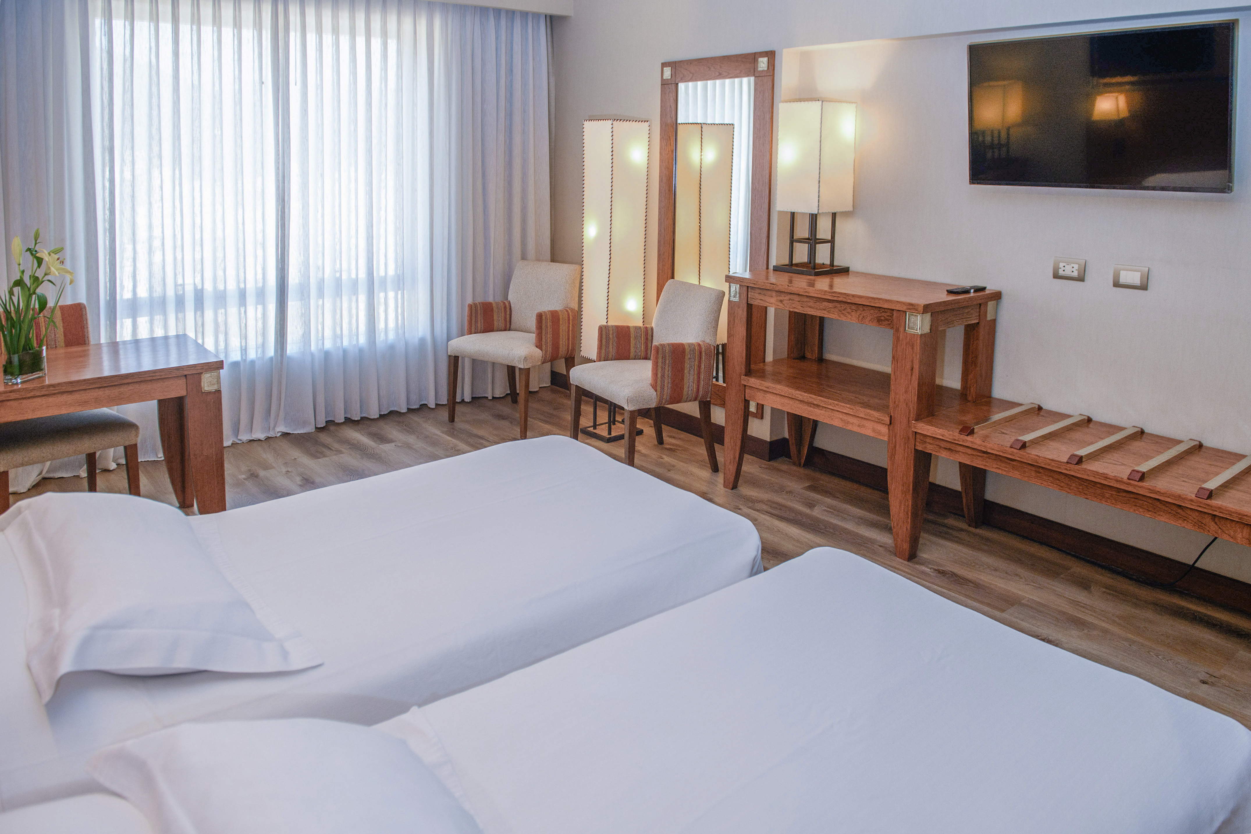 Modern hotel room with twin beds, wooden furniture, and sheer window curtains.