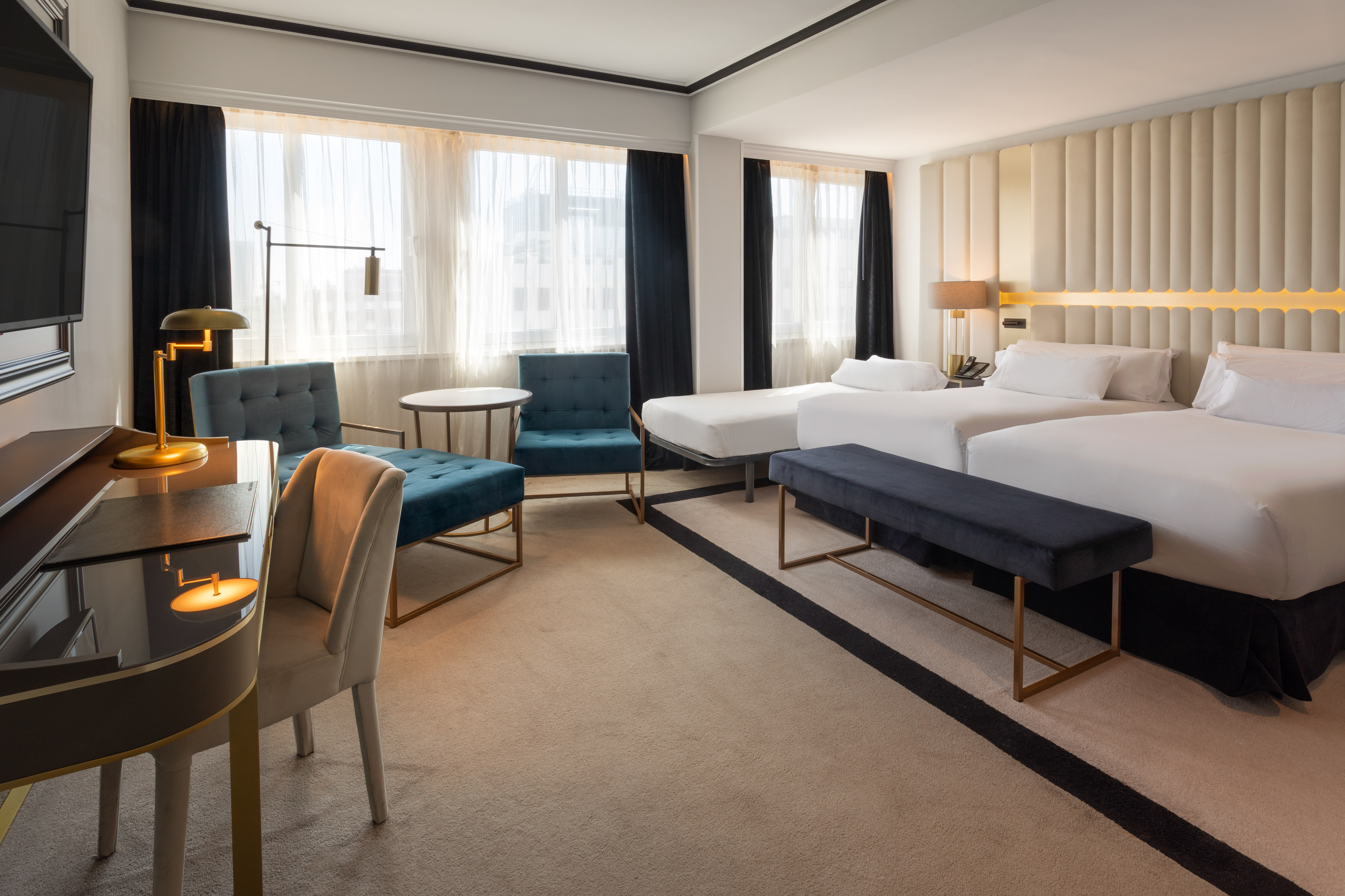 Elegant hotel room with twin beds, modern desk, and plush blue seating area.