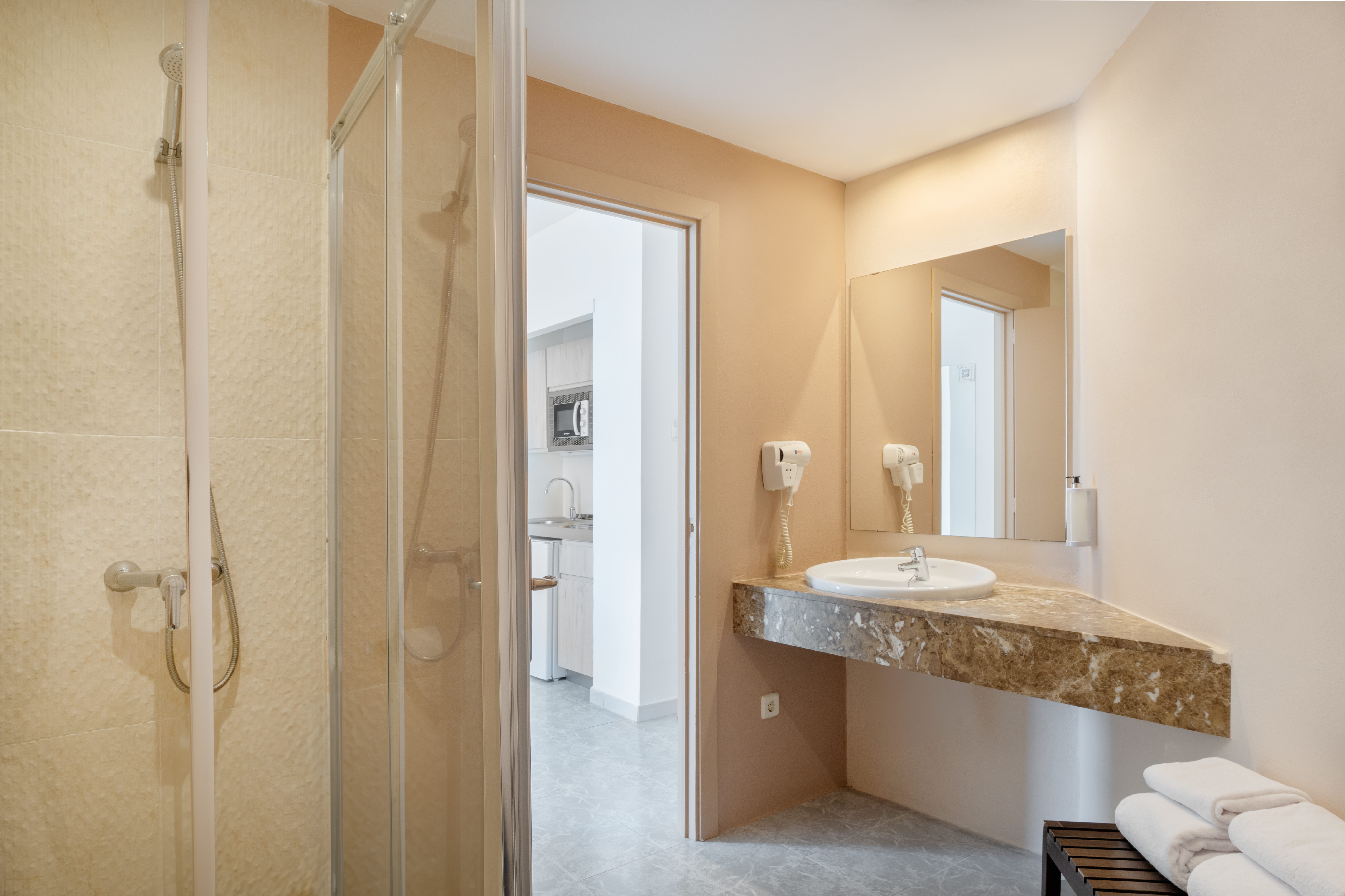 Hotel bathroom with shower, marble vanity, sink, mirror, and kitchen view.