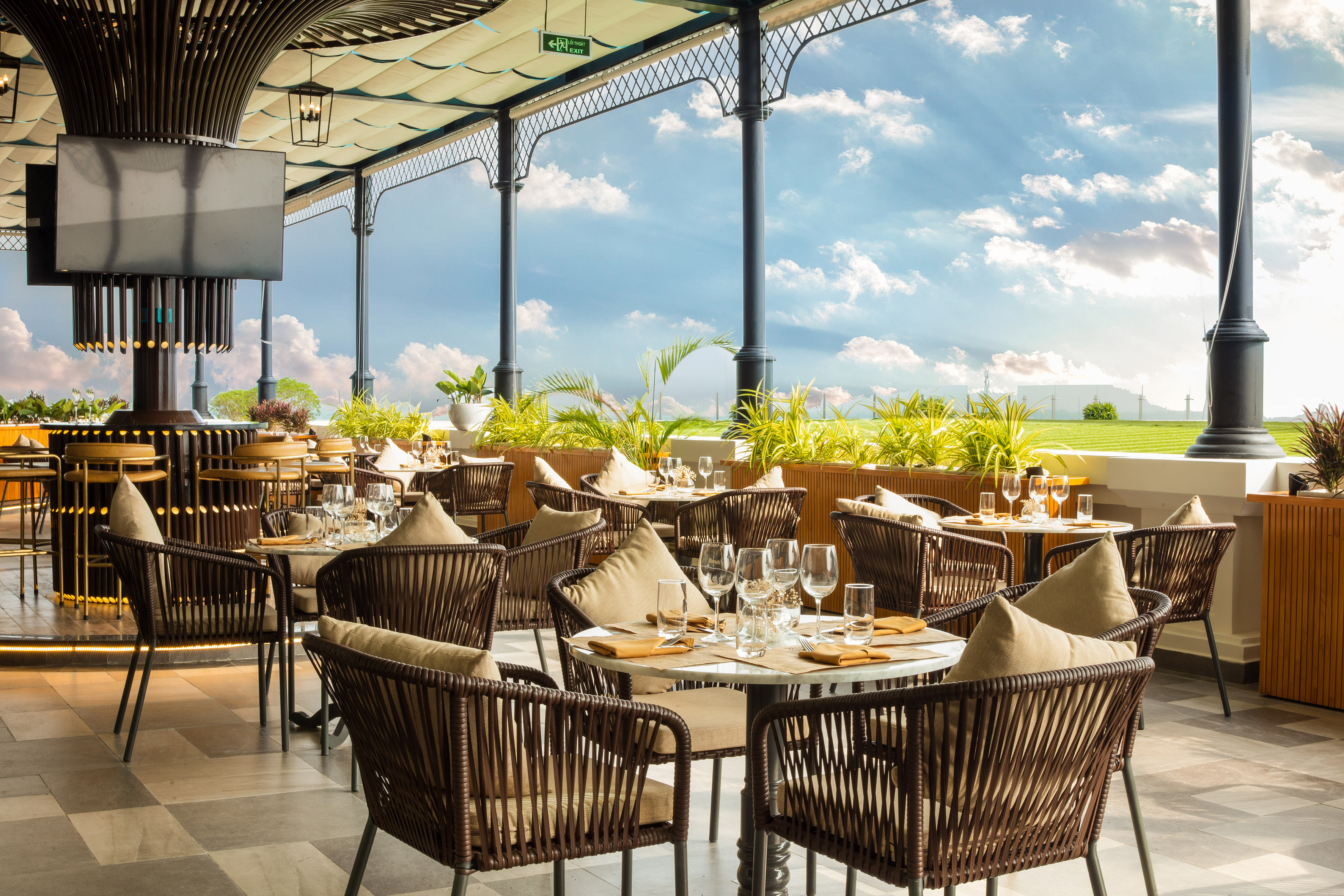 Outdoor restaurant patio with tables, chairs, lush plants, and bright blue sky.