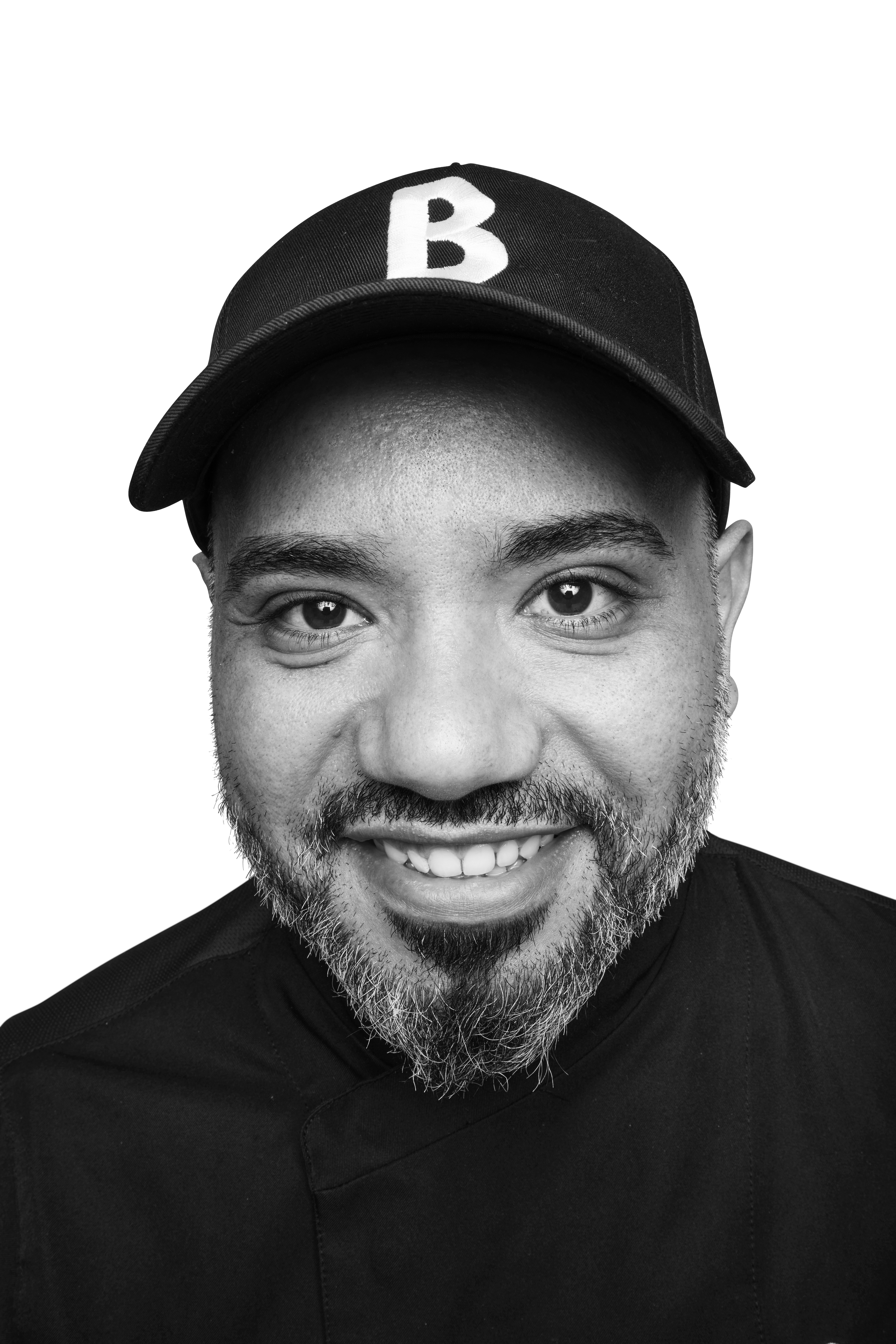 Smiling man with beard, wearing black cap with 'B' logo, black and white portrait.