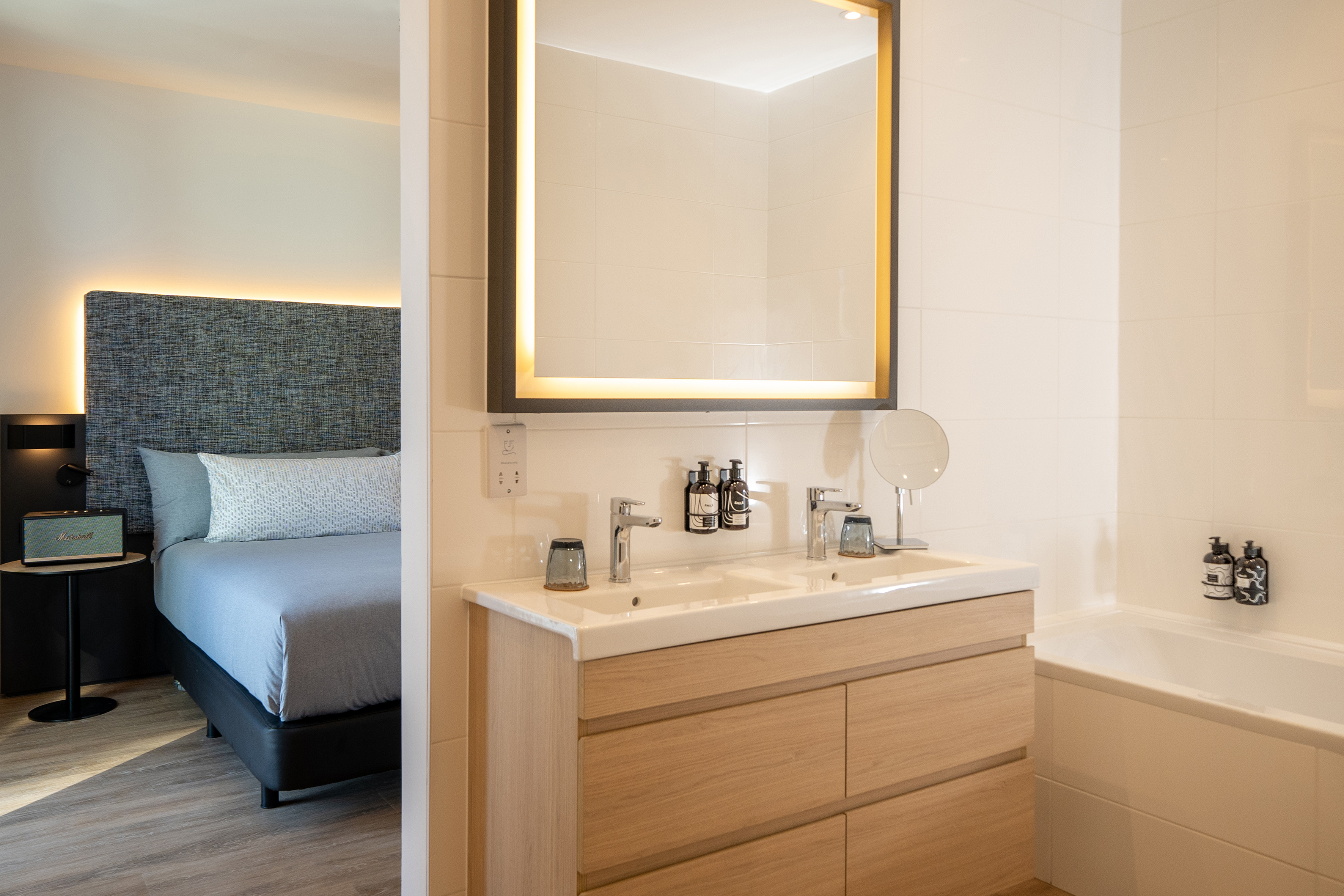 Modern hotel room: bathroom with double vanity, backlit mirror, and bed.