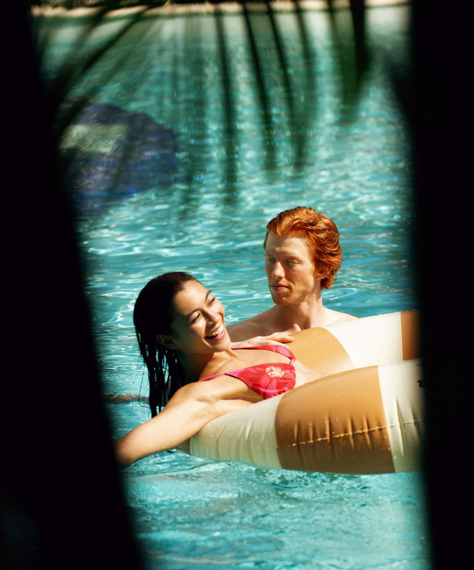 Smiling woman on inflatable pool float, man with red hair in blue water.