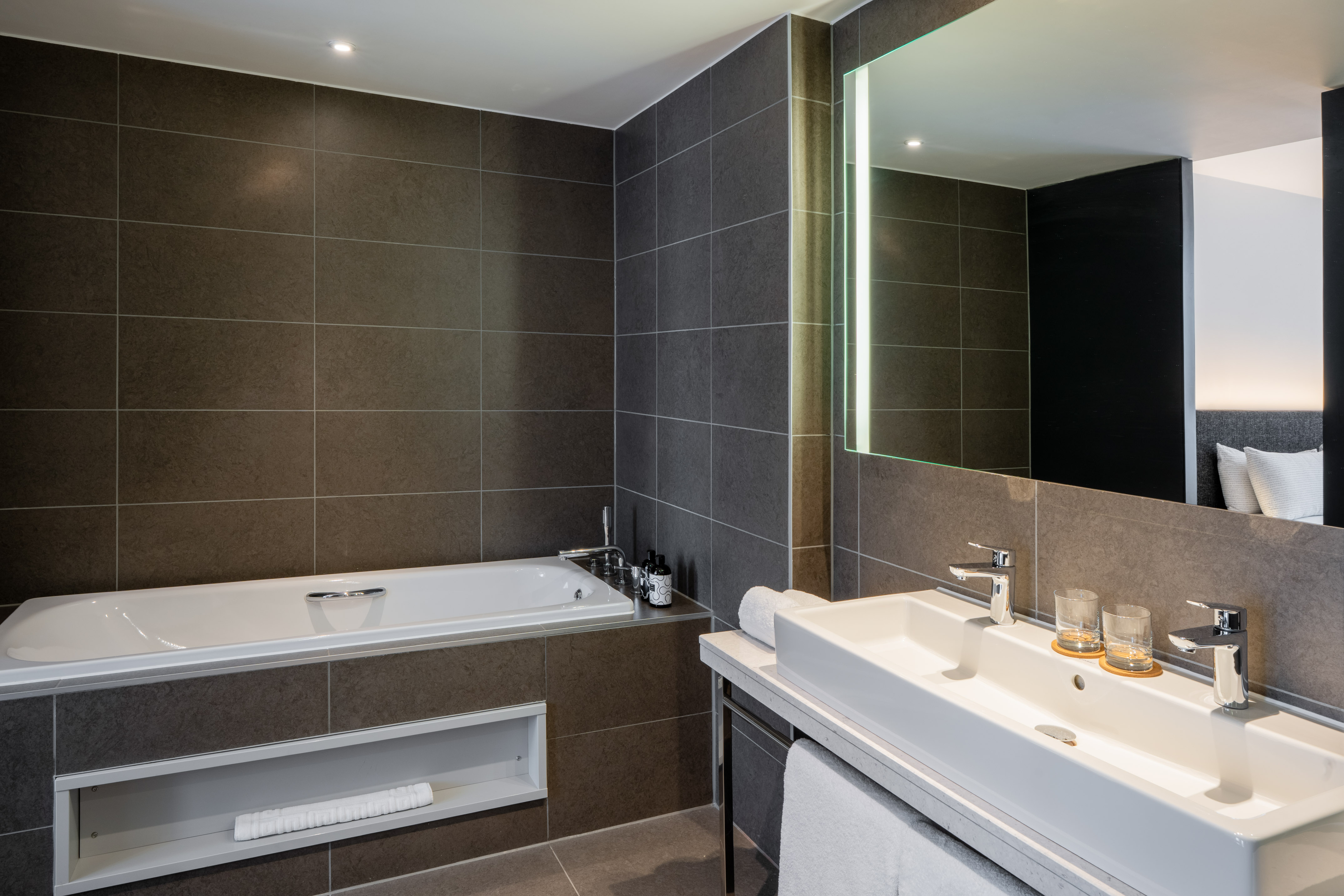 Modern hotel bathroom with dark grey tiles, bathtub, double sink, and illuminated mirror.