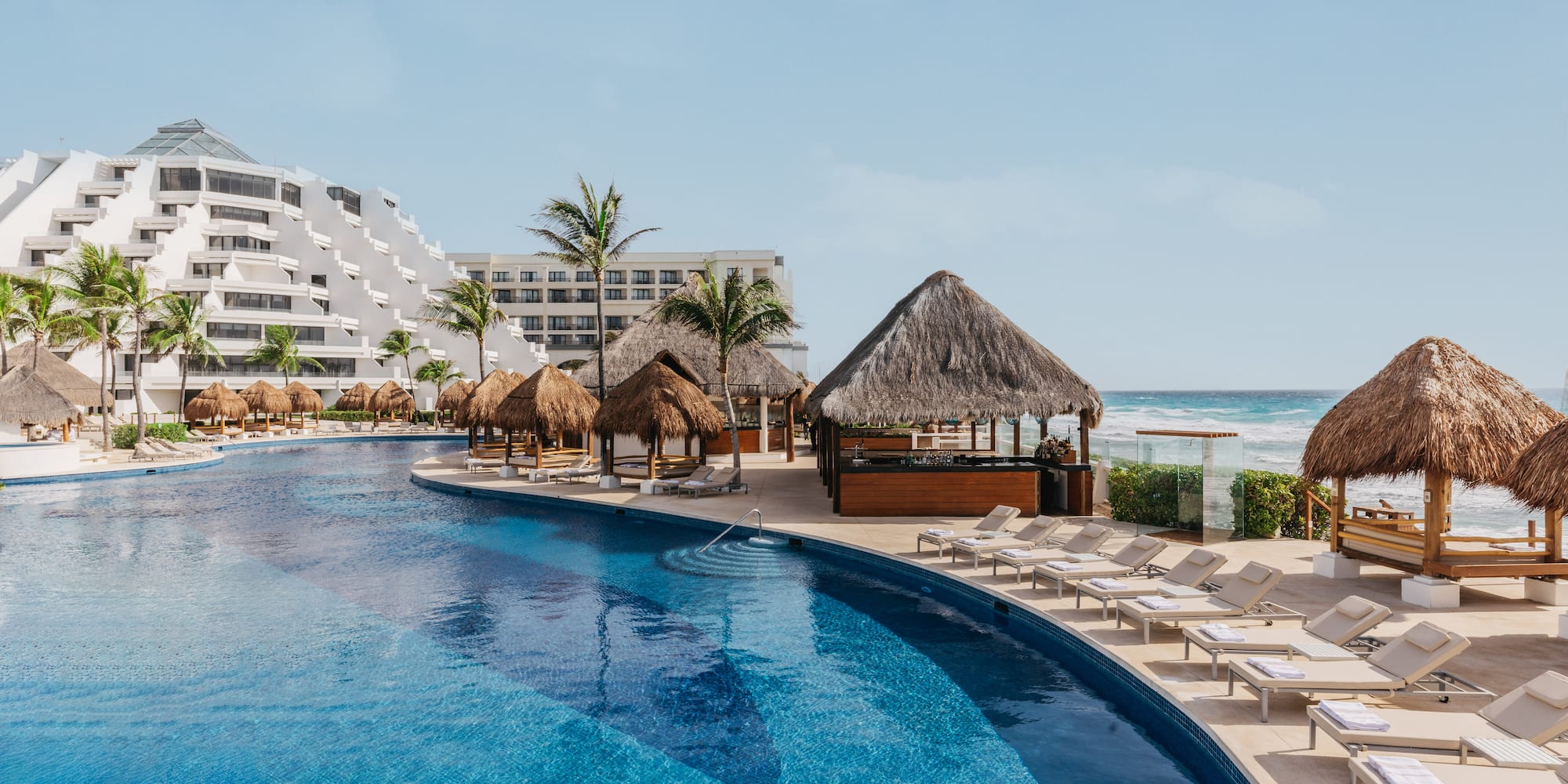 Cancun resort pool with beach, palapas, palm trees, and tiered hotel.