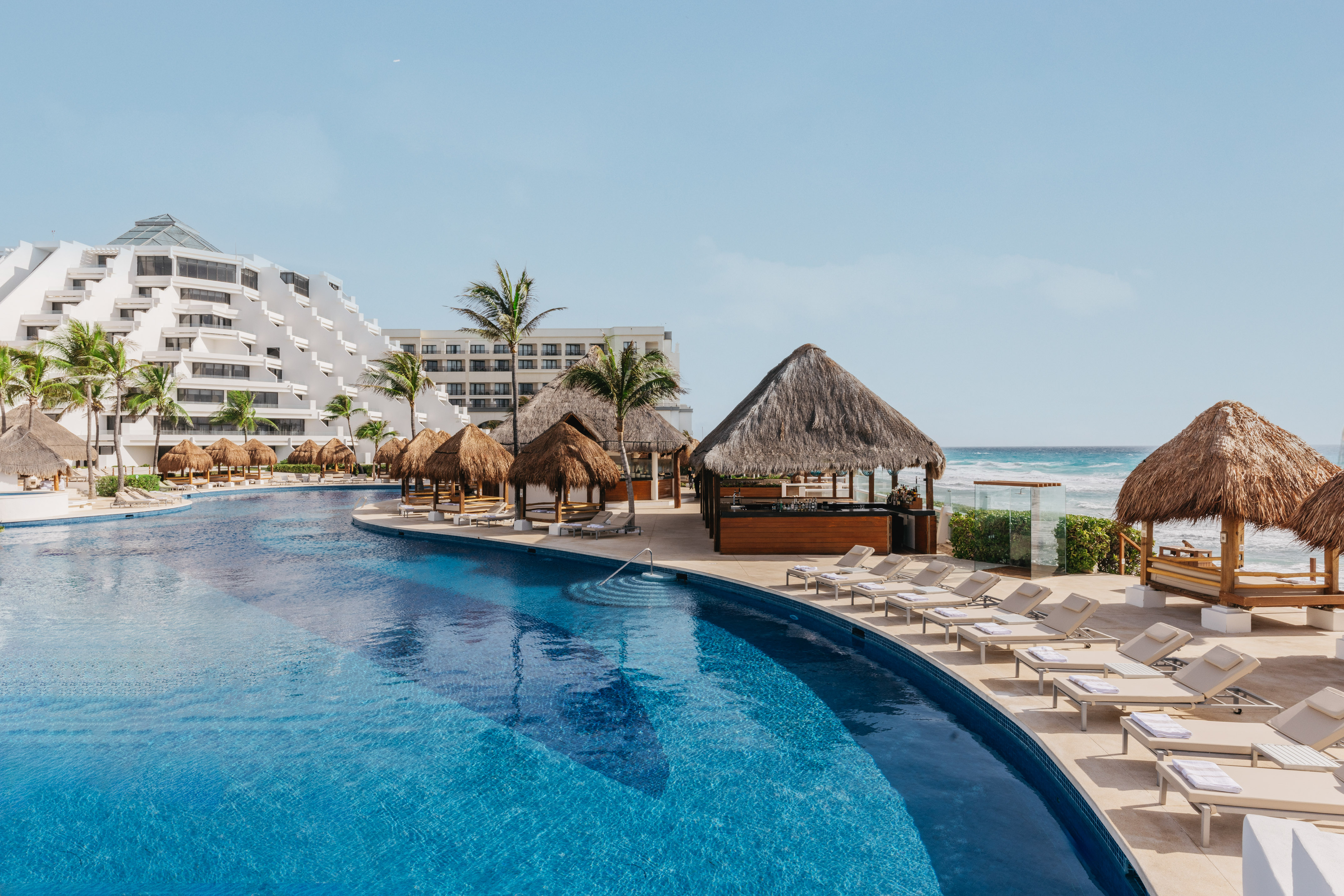 Cancun resort pool with beach, palapas, palm trees, and tiered hotel.