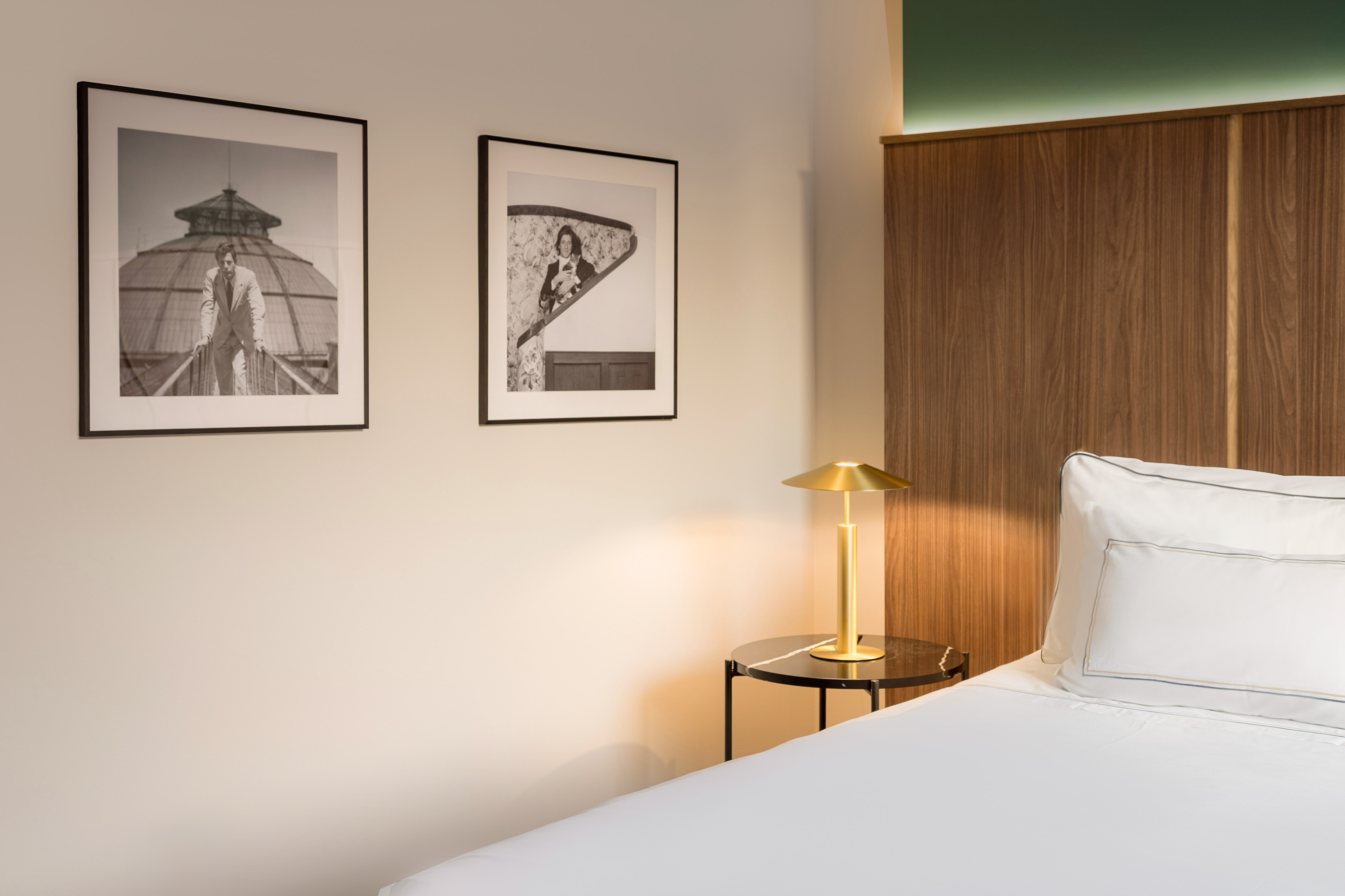 Modern hotel room: white bed, wooden headboard, golden lamp, framed art.