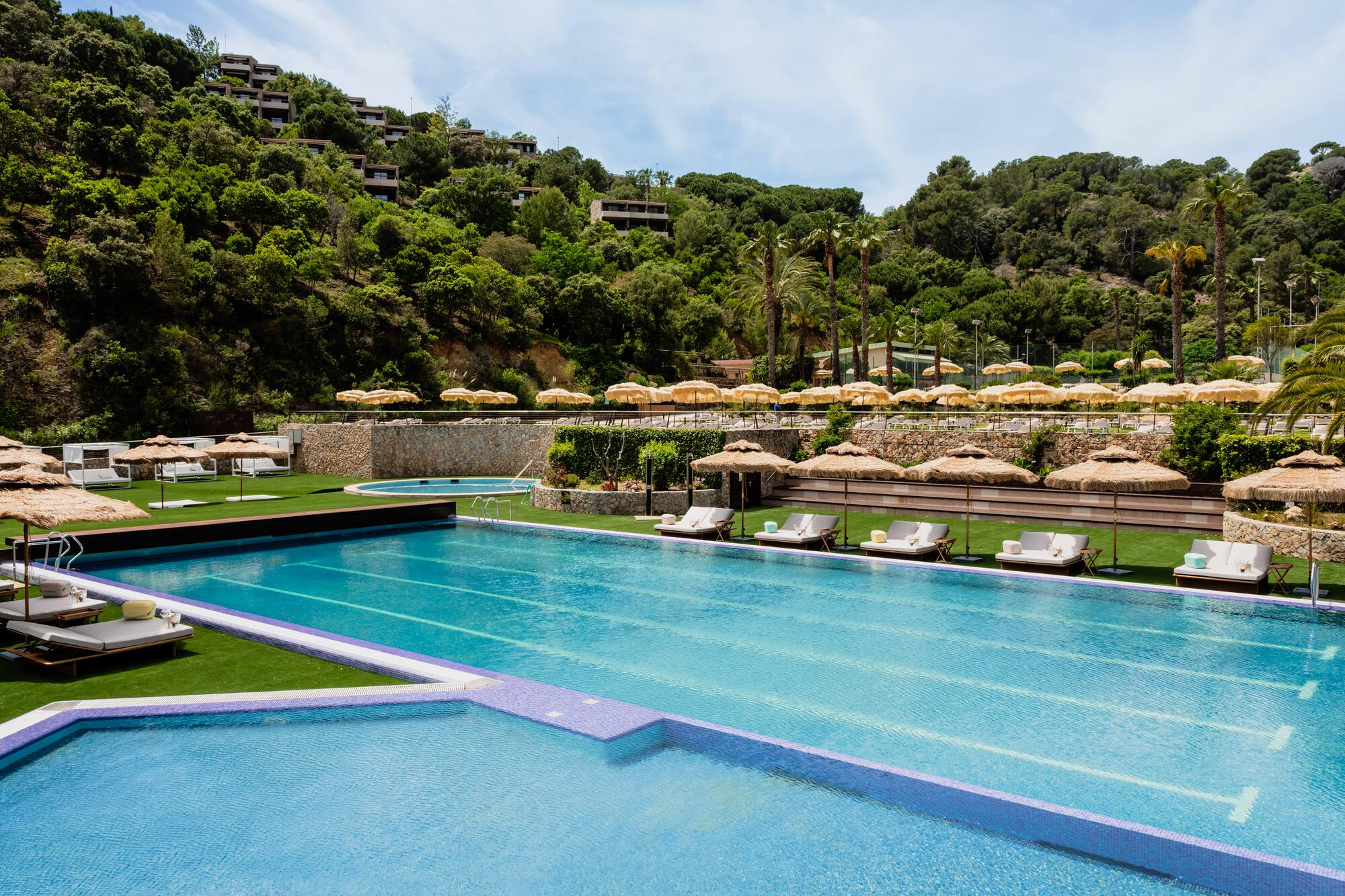 Resort swimming pool with lounge chairs, straw umbrellas, and hillside hotel.