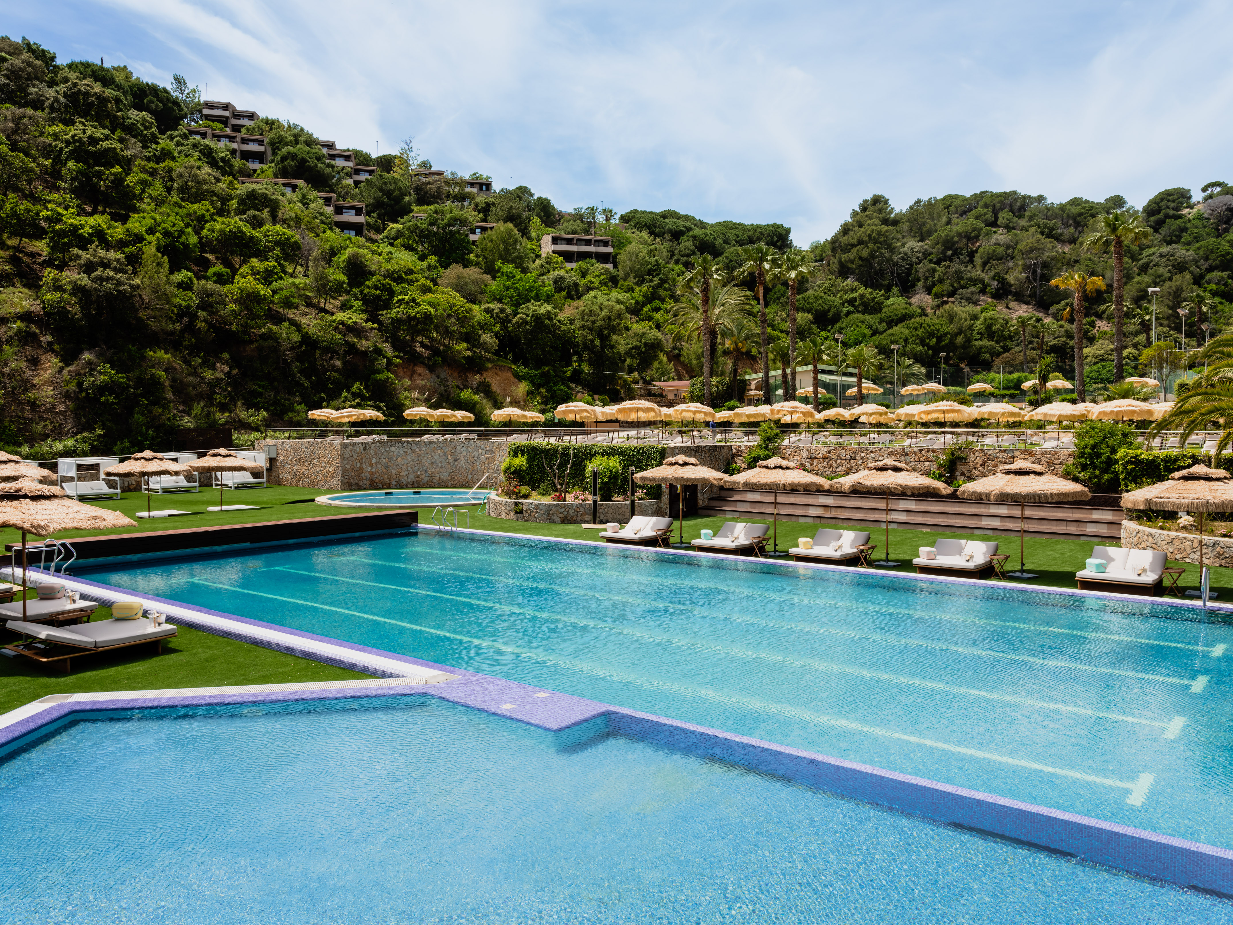 Resort swimming pool with lounge chairs, straw umbrellas, and hillside hotel.