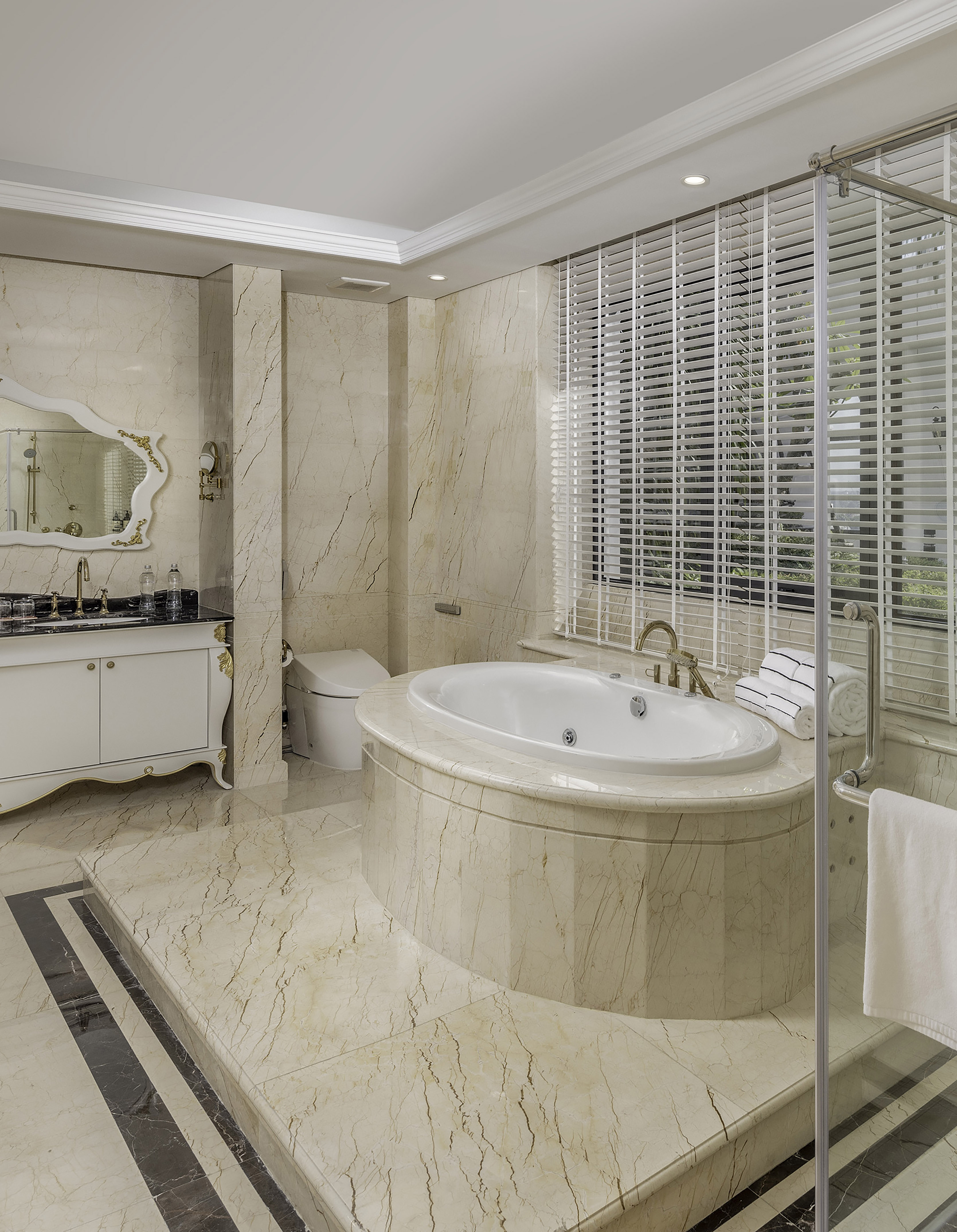 Elegant marble bathroom with oval soaking tub, ornate vanity, and gold fixtures.