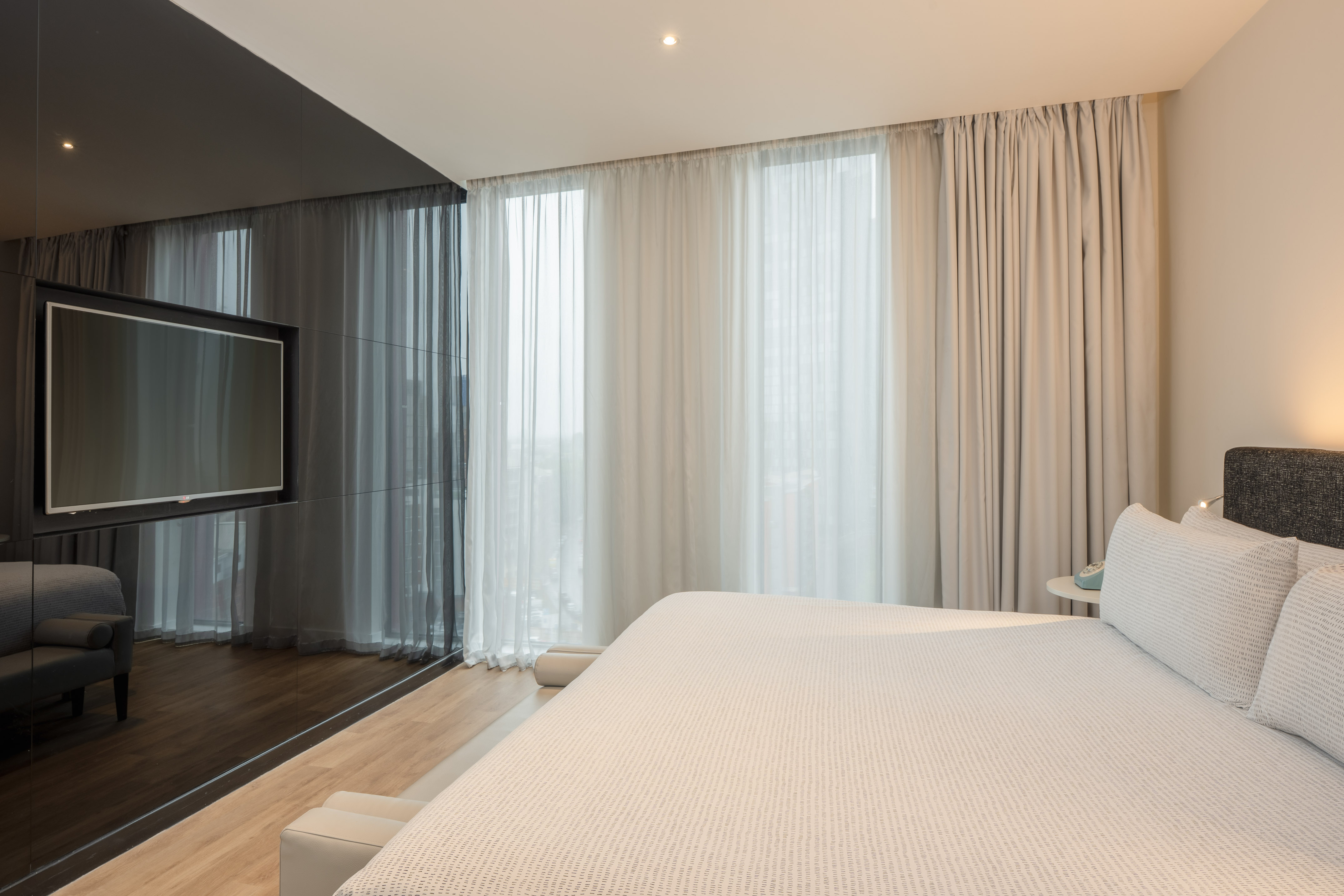 Modern hotel bedroom with king bed, wall TV, sheer curtains, and wood floor.