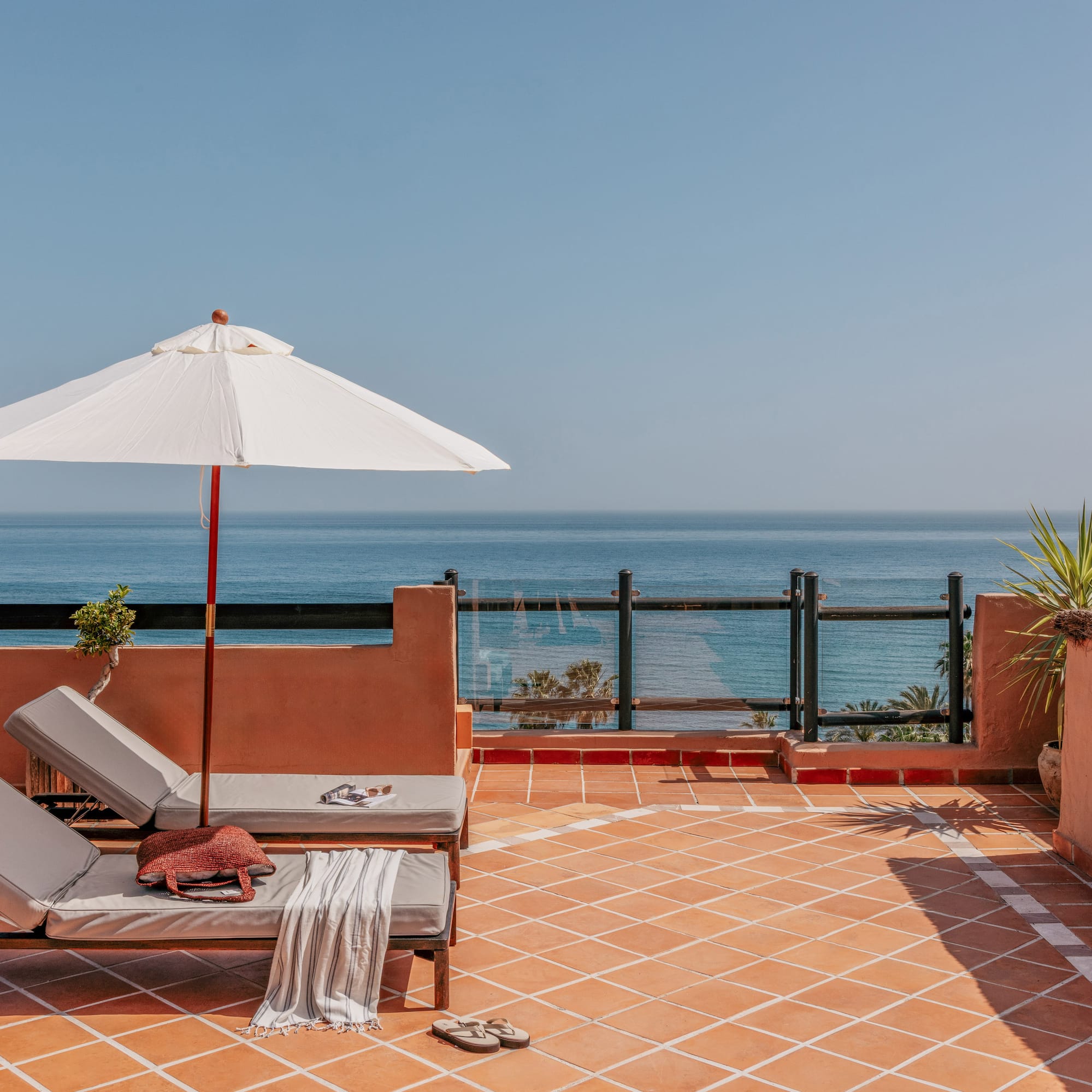 Terracotta rooftop terrace with ocean view, two sun loungers, and white sun umbrella.