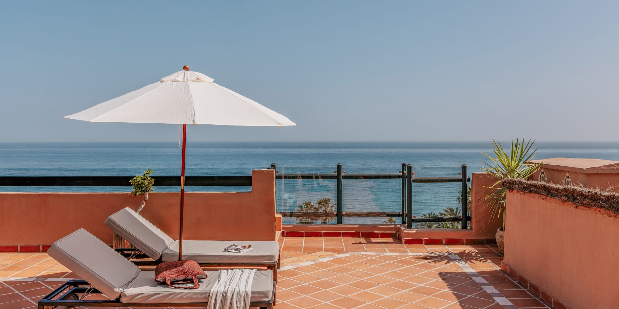 Terracotta rooftop terrace with ocean view, two sun loungers, and white sun umbrella.