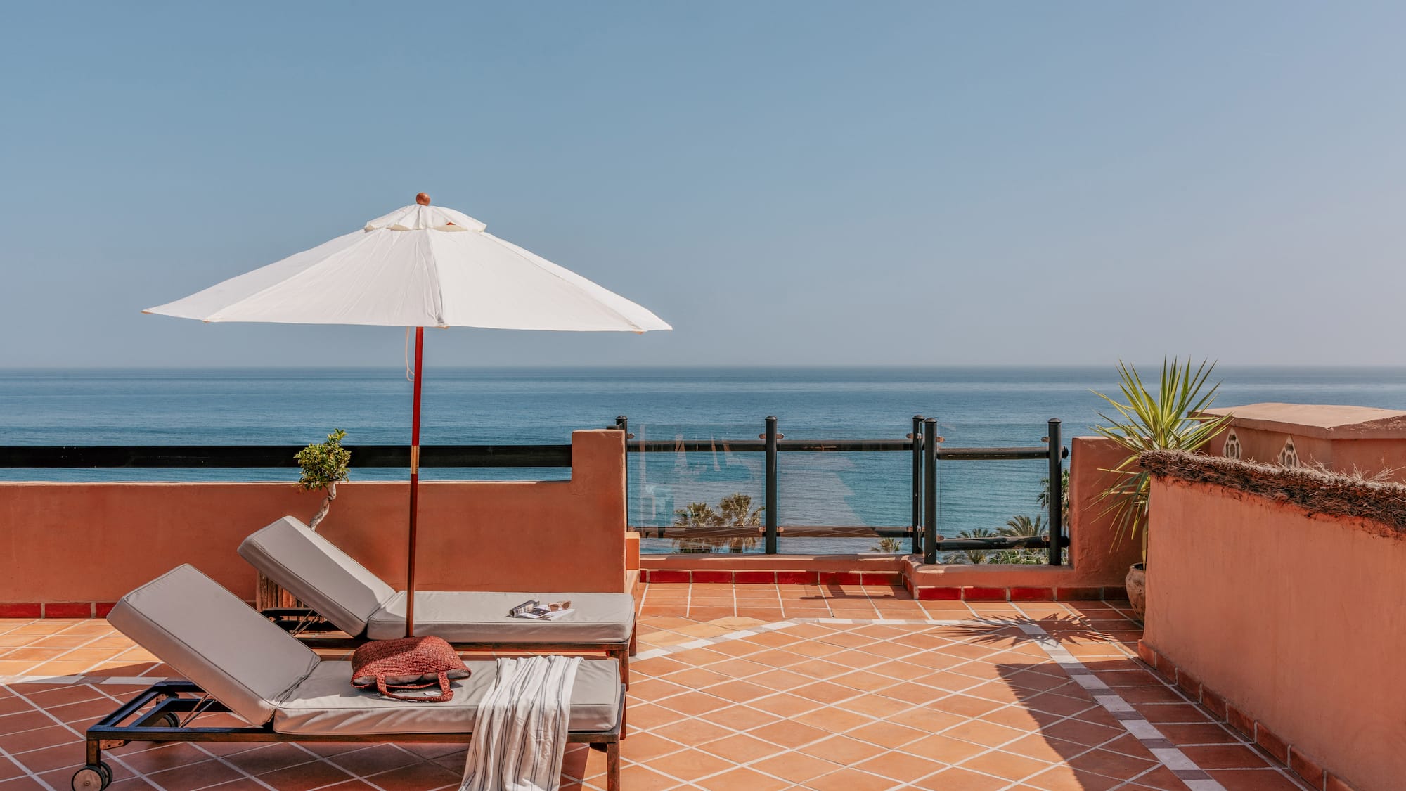 Terracotta rooftop terrace with ocean view, two sun loungers, and white sun umbrella.