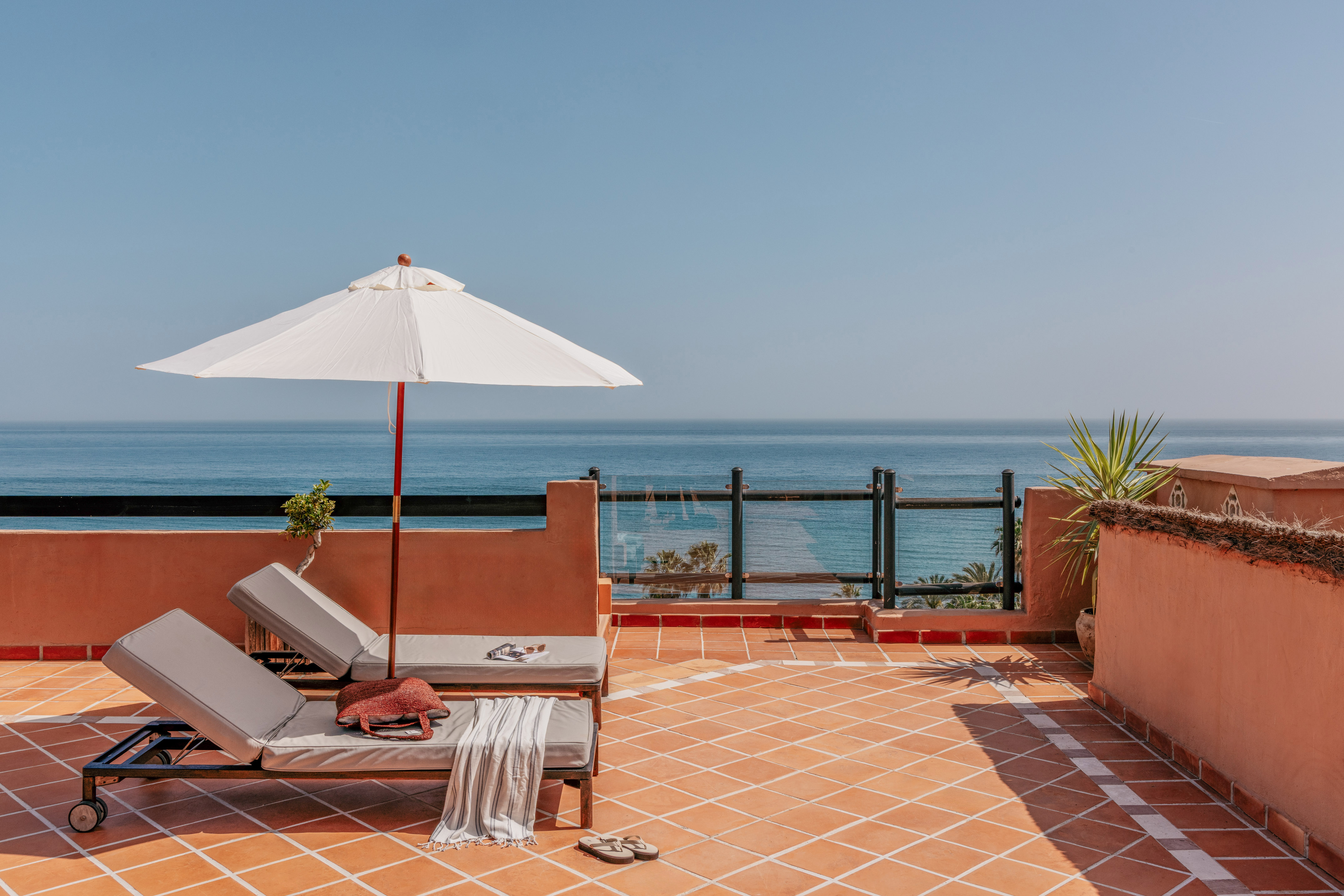 Terracotta rooftop terrace with ocean view, two sun loungers, and white sun umbrella.