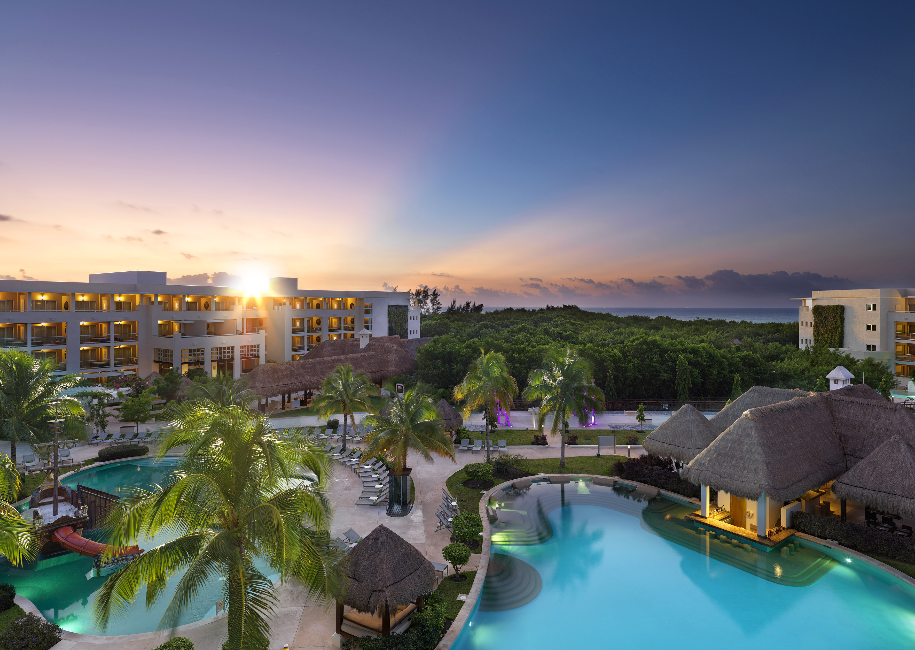 a pool with palm trees and a building with a beach and a sunset