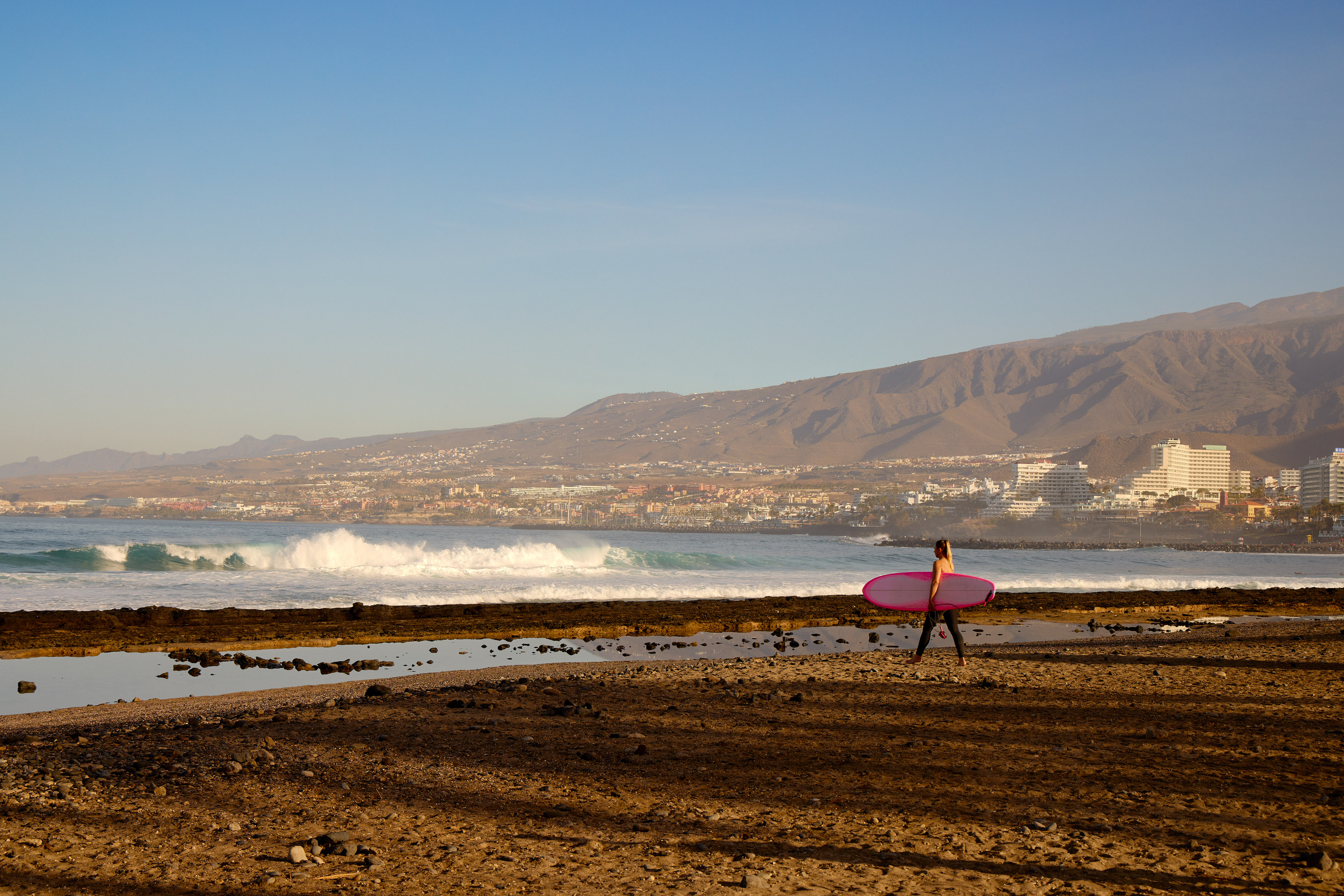 Surfer with pink board walking rocky beach, ocean waves, coastal city, mountains.