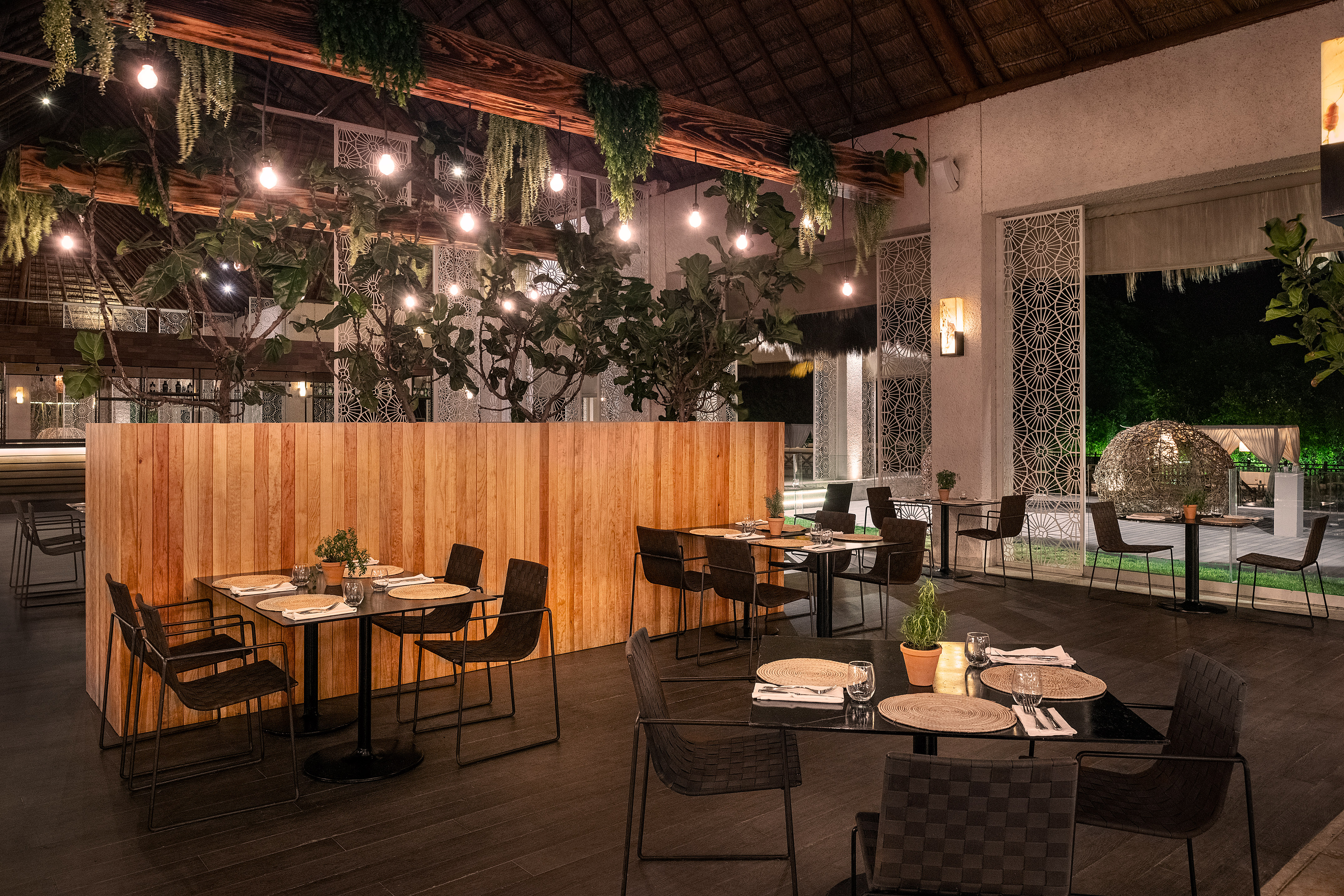Tropical restaurant dining area at night, warm lighting, plants, and thatch roof.