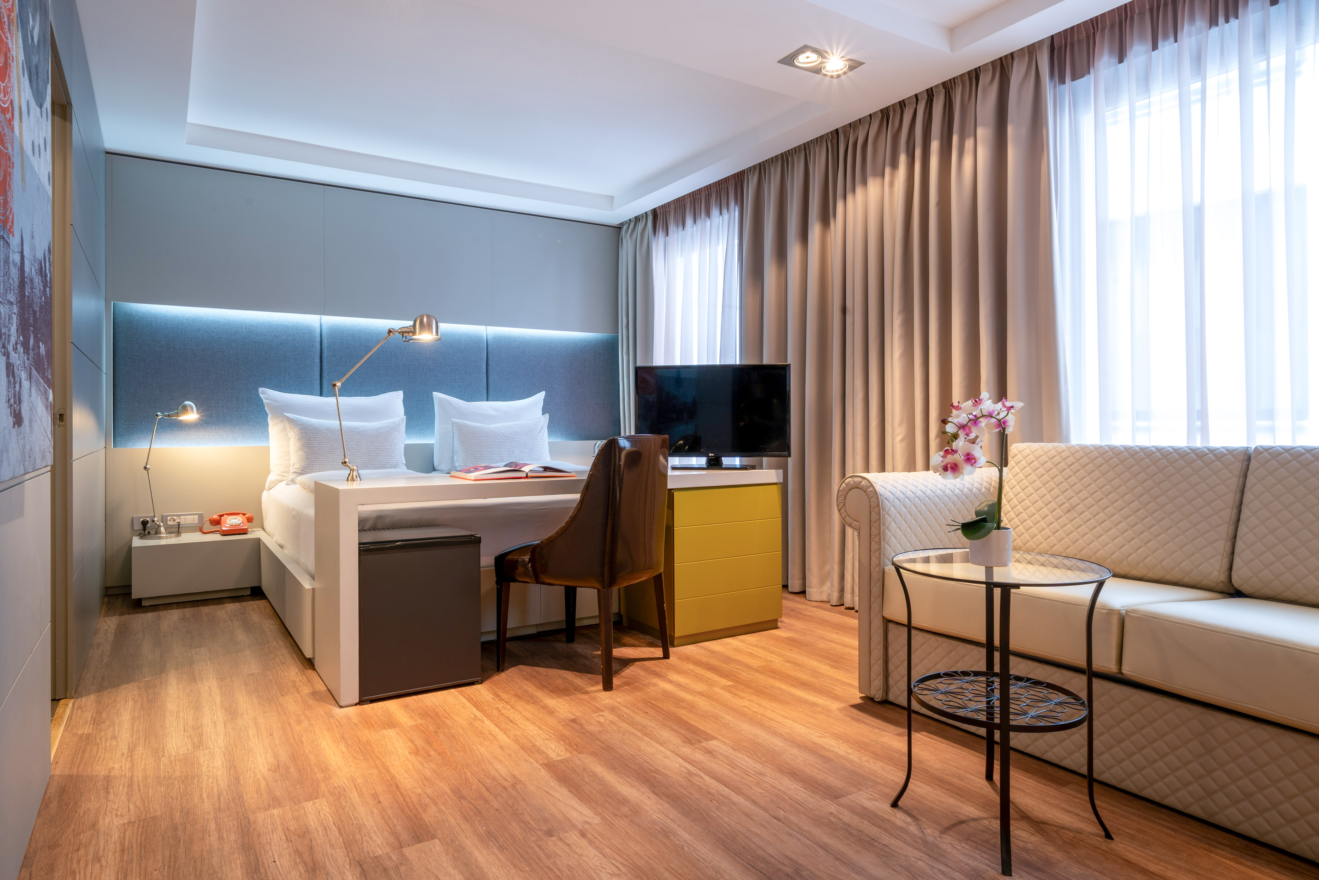 Contemporary hotel suite with bed, desk, sofa, TV, and wood floor.