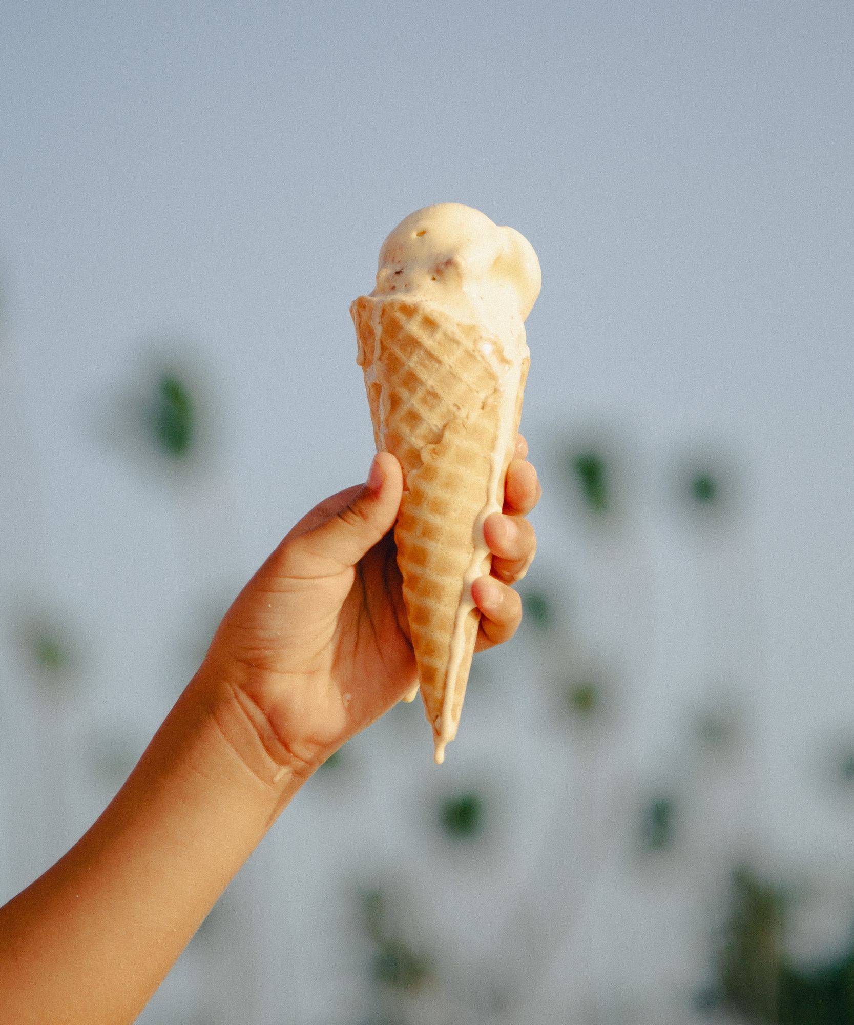 Hand holding a melting ice cream cone against a blurry blue background.