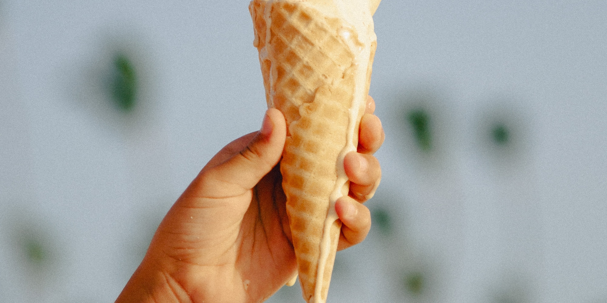 Hand holds melting ice cream in a waffle cone against blue sky.
