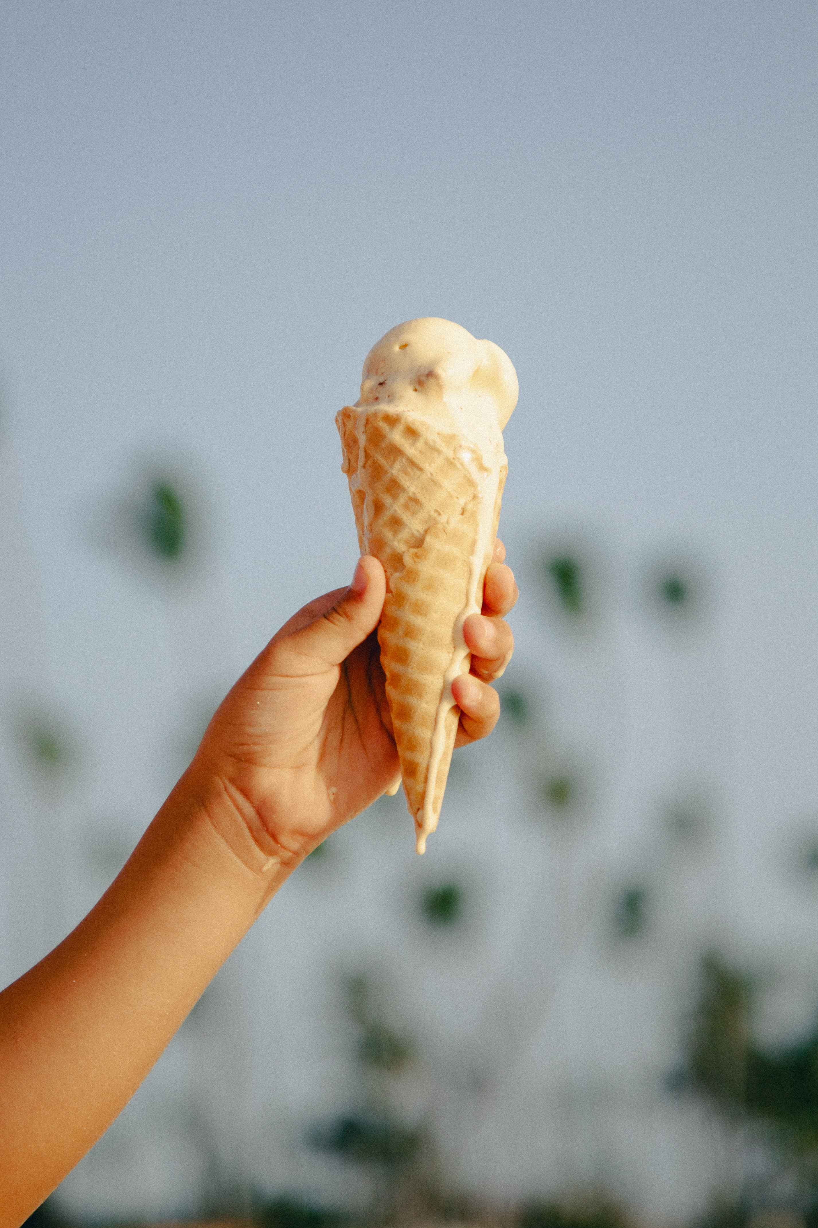 Hand holding melting ice cream in a waffle cone against a blurry sky.