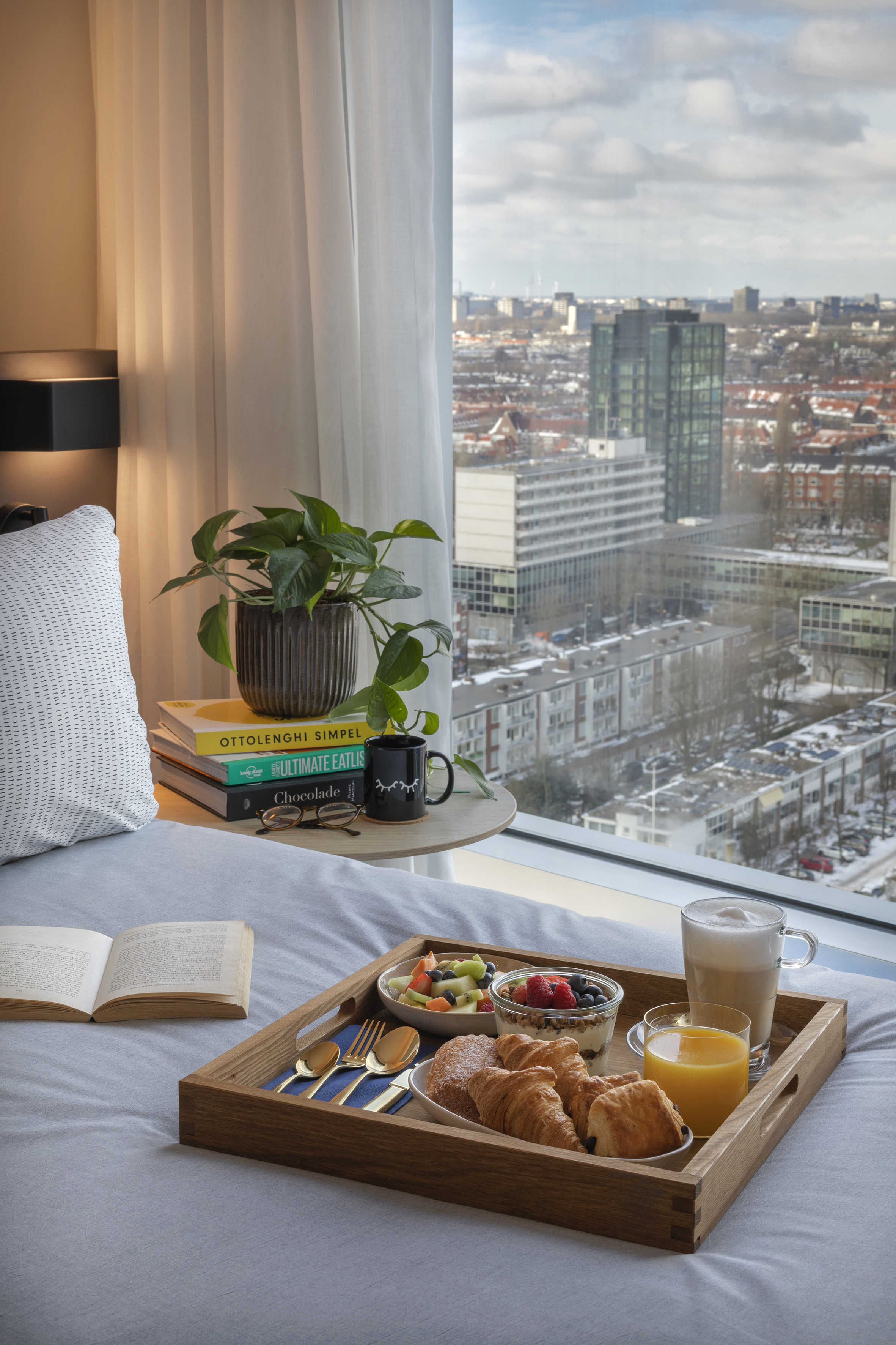 Hotel room with breakfast in bed, city view from window, croissants, coffee.