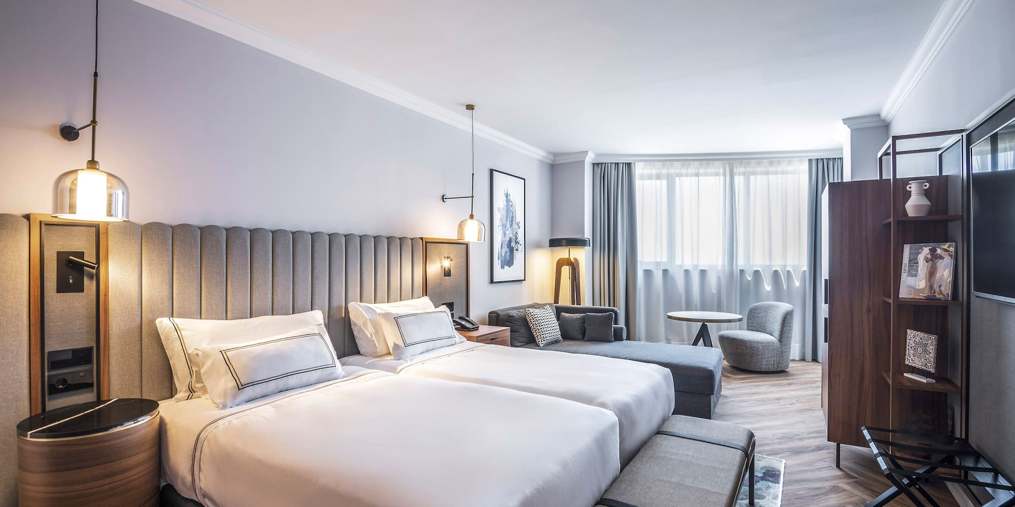 Habitación de hotel moderna con dos camas, sofá y ventanal luminoso.