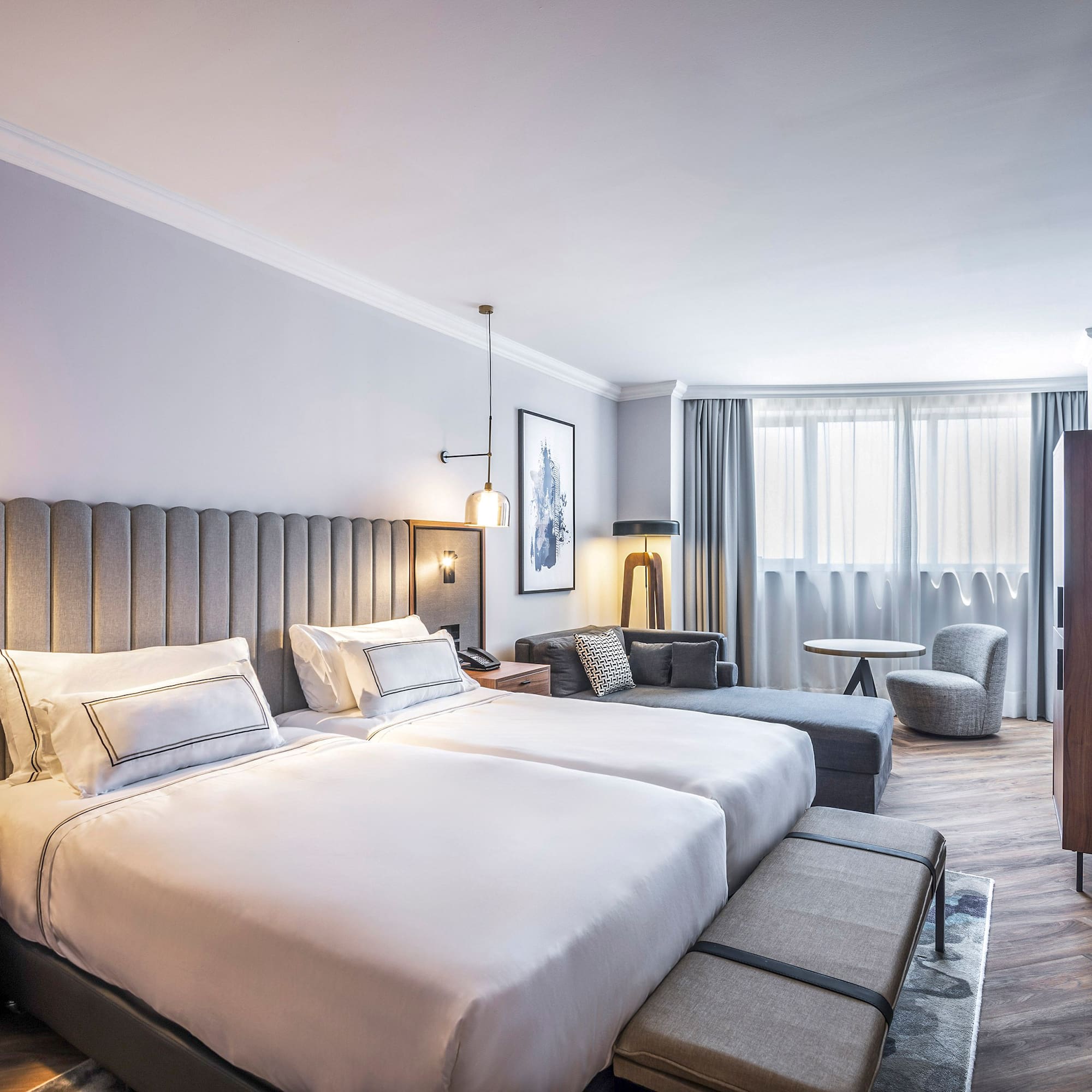 Habitación de hotel moderna con dos camas, sofá y ventanal luminoso.