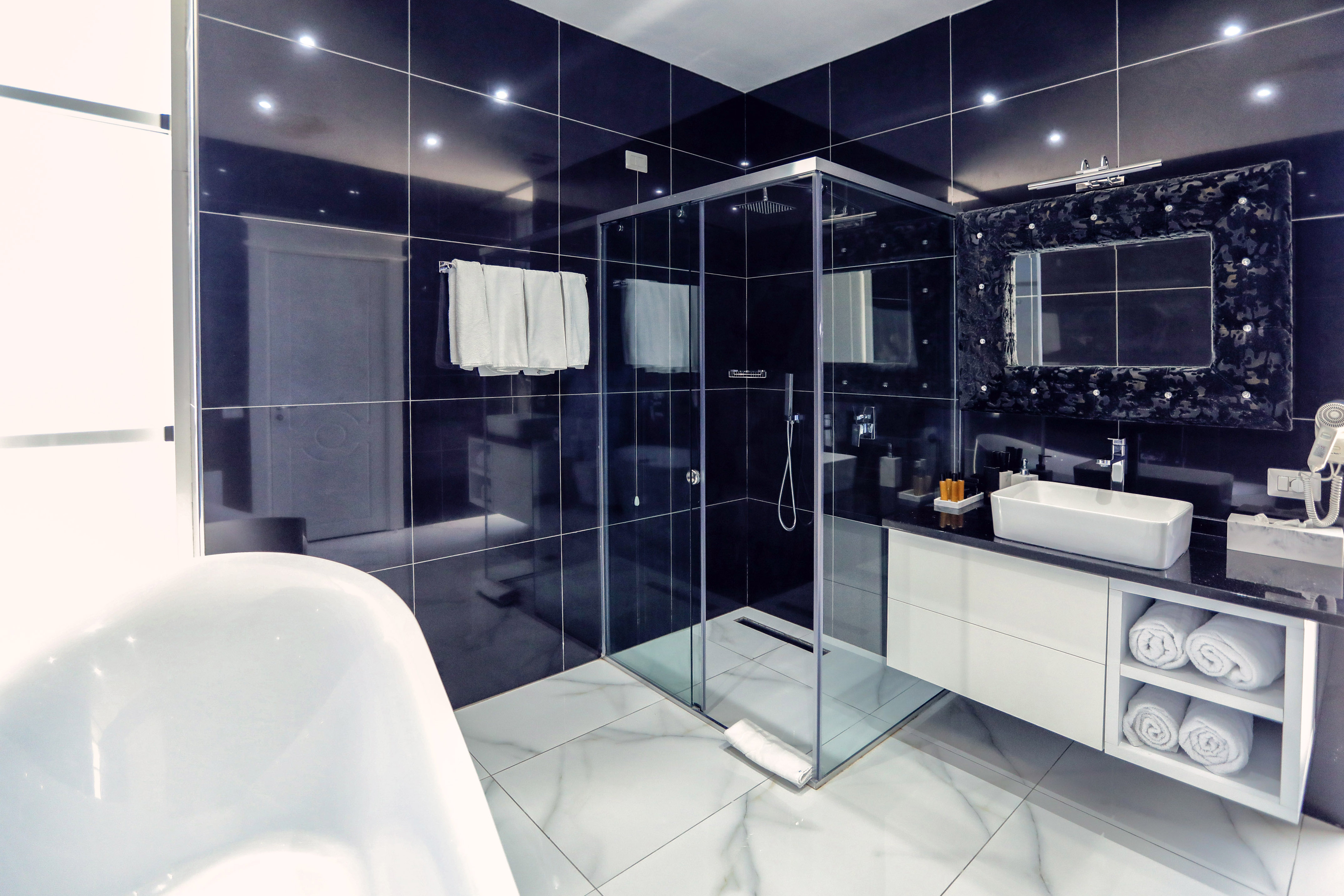 Modern black tiled bathroom with glass shower, white tub, and vanity.