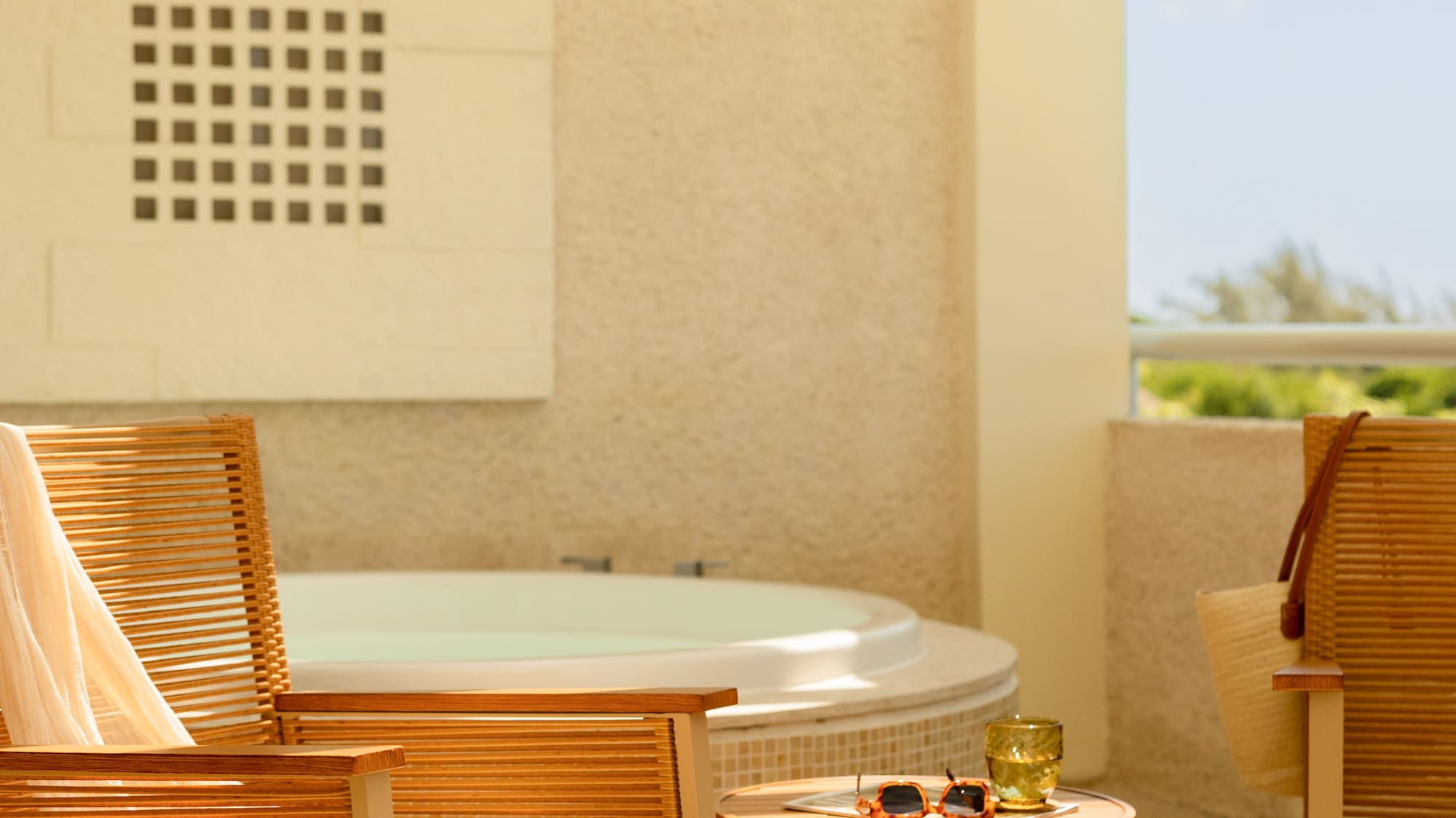 Sunny hotel balcony with hot tub, wooden rocking chair, and wicker table.