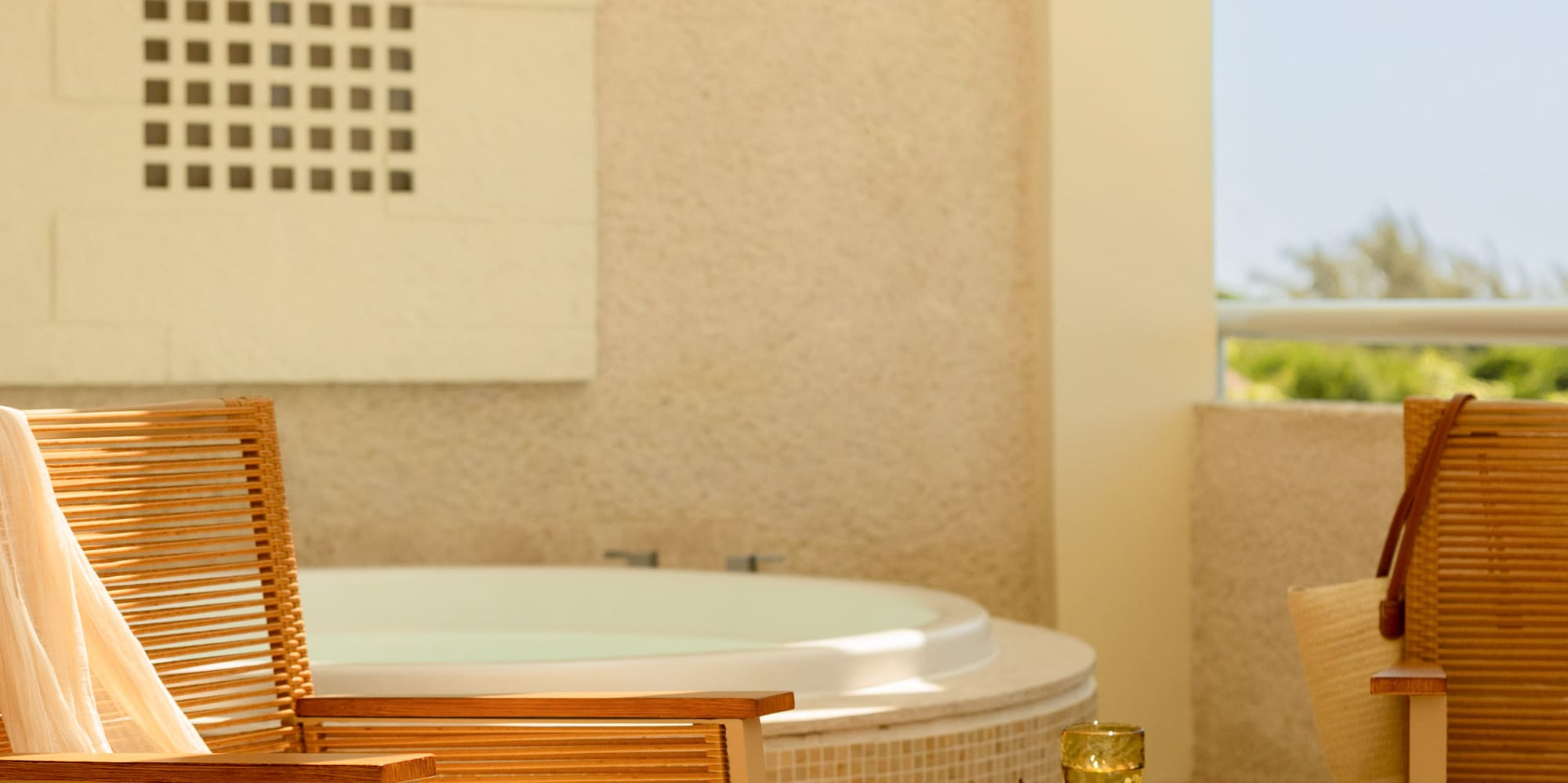 Sunny hotel balcony with hot tub, wooden rocking chair, and wicker table.