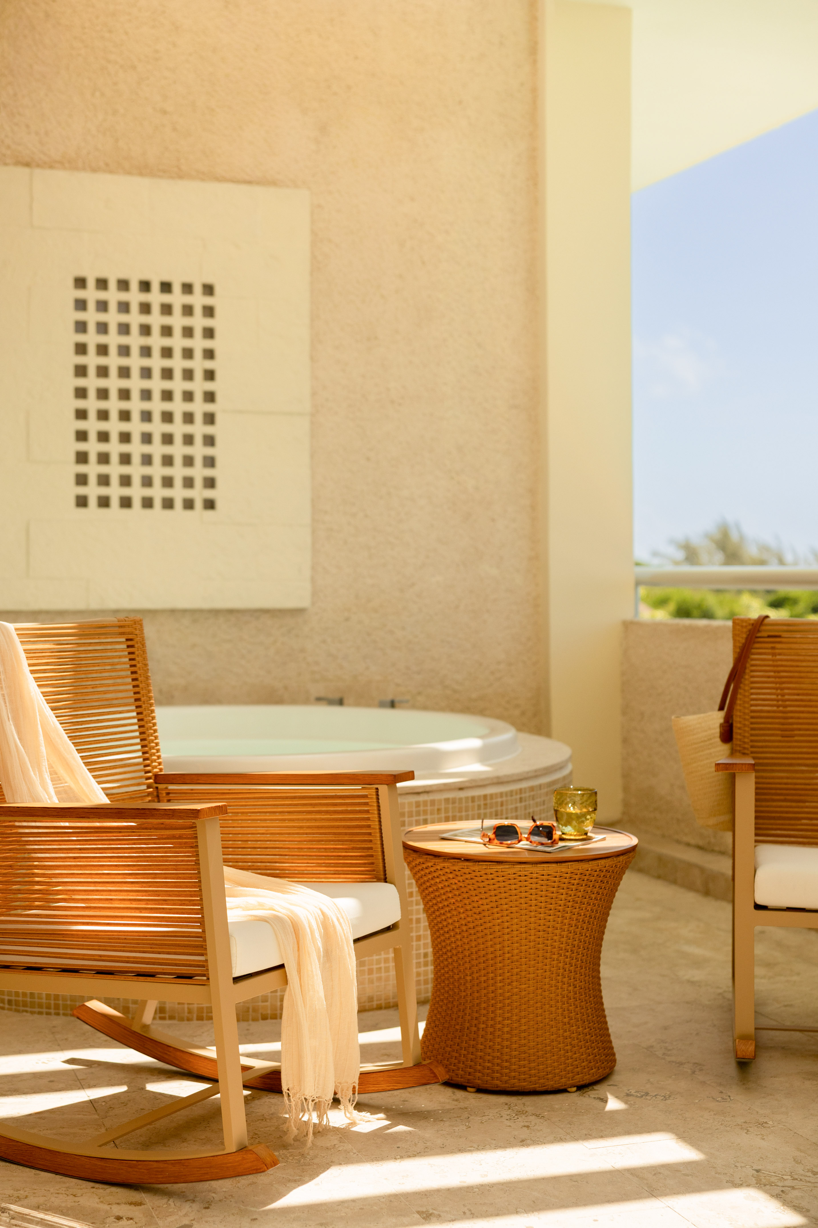 Sunny hotel balcony with hot tub, wooden rocking chair, and wicker table.
