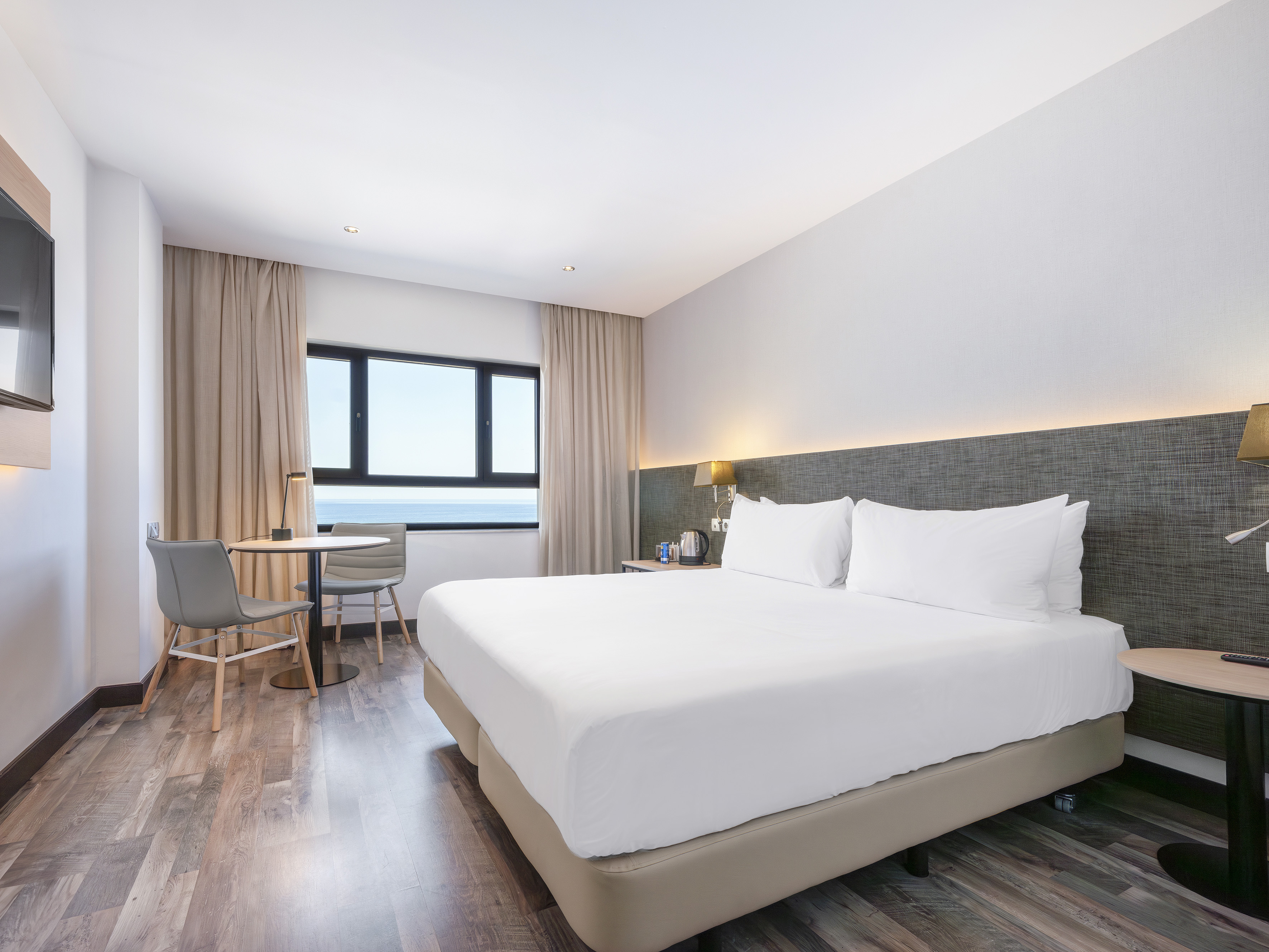 Bright hotel room with king bed, sea view window, and modern decor.