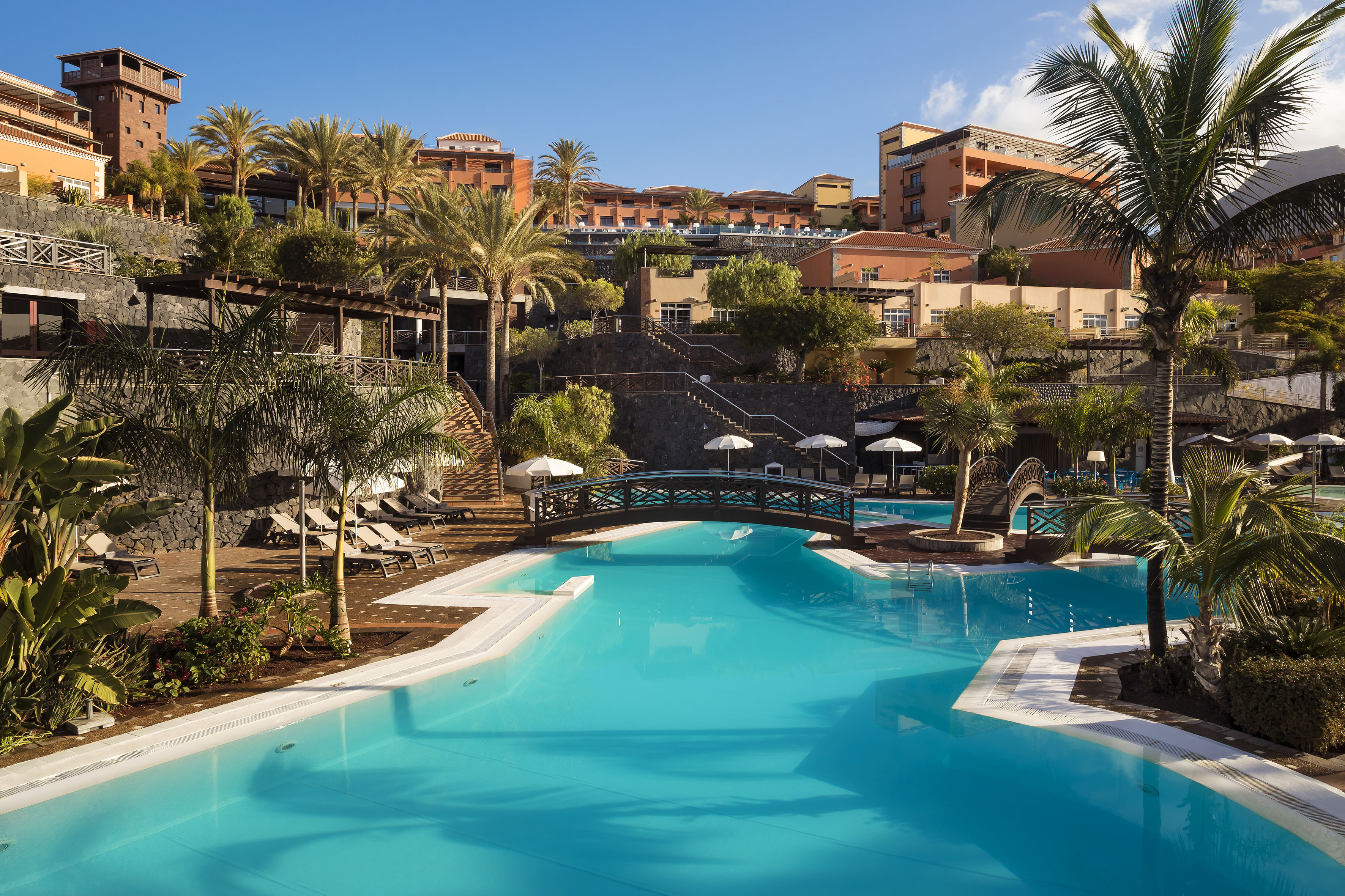 Luxury resort swimming pool with bridge, palm trees, and terraced hotel buildings.