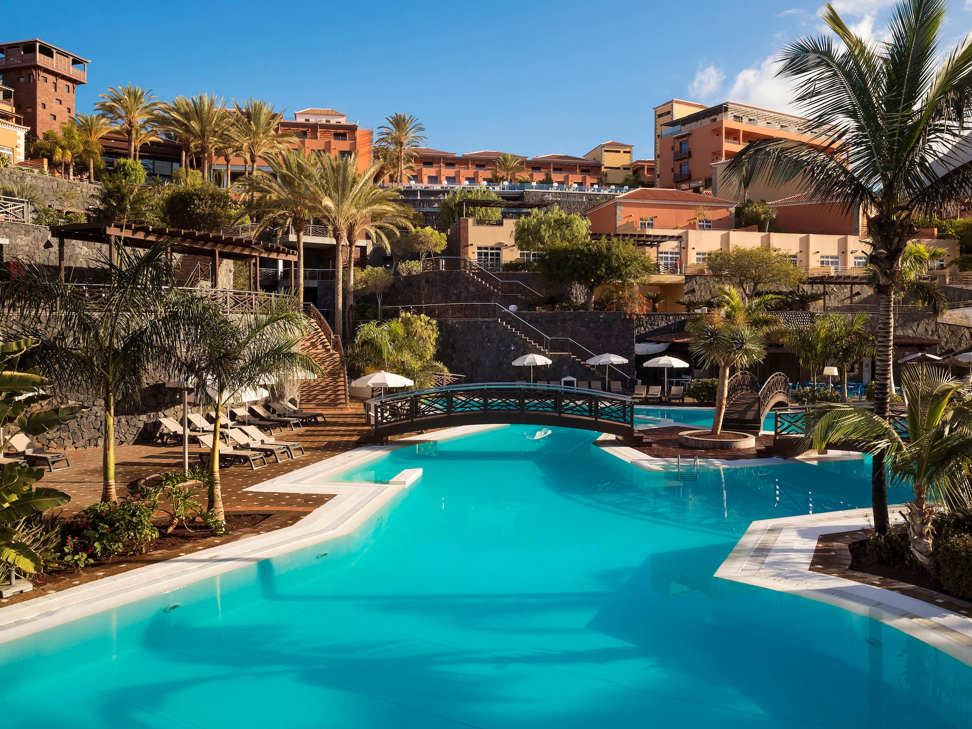 Luxury resort swimming pool with bridge, palm trees, and terraced hotel buildings.