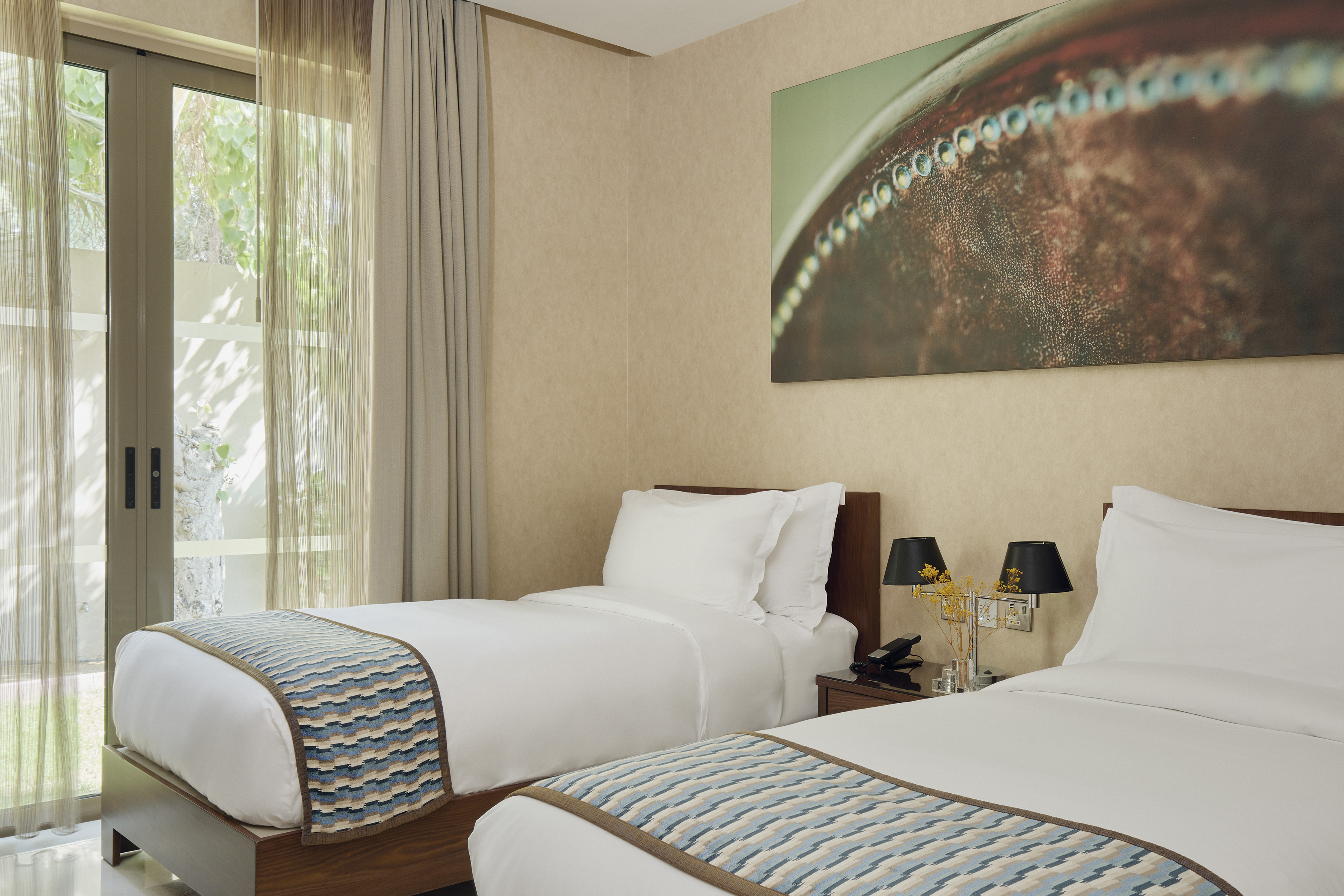 Twin bed hotel room with patterned runners, bedside lamps, and large window view.