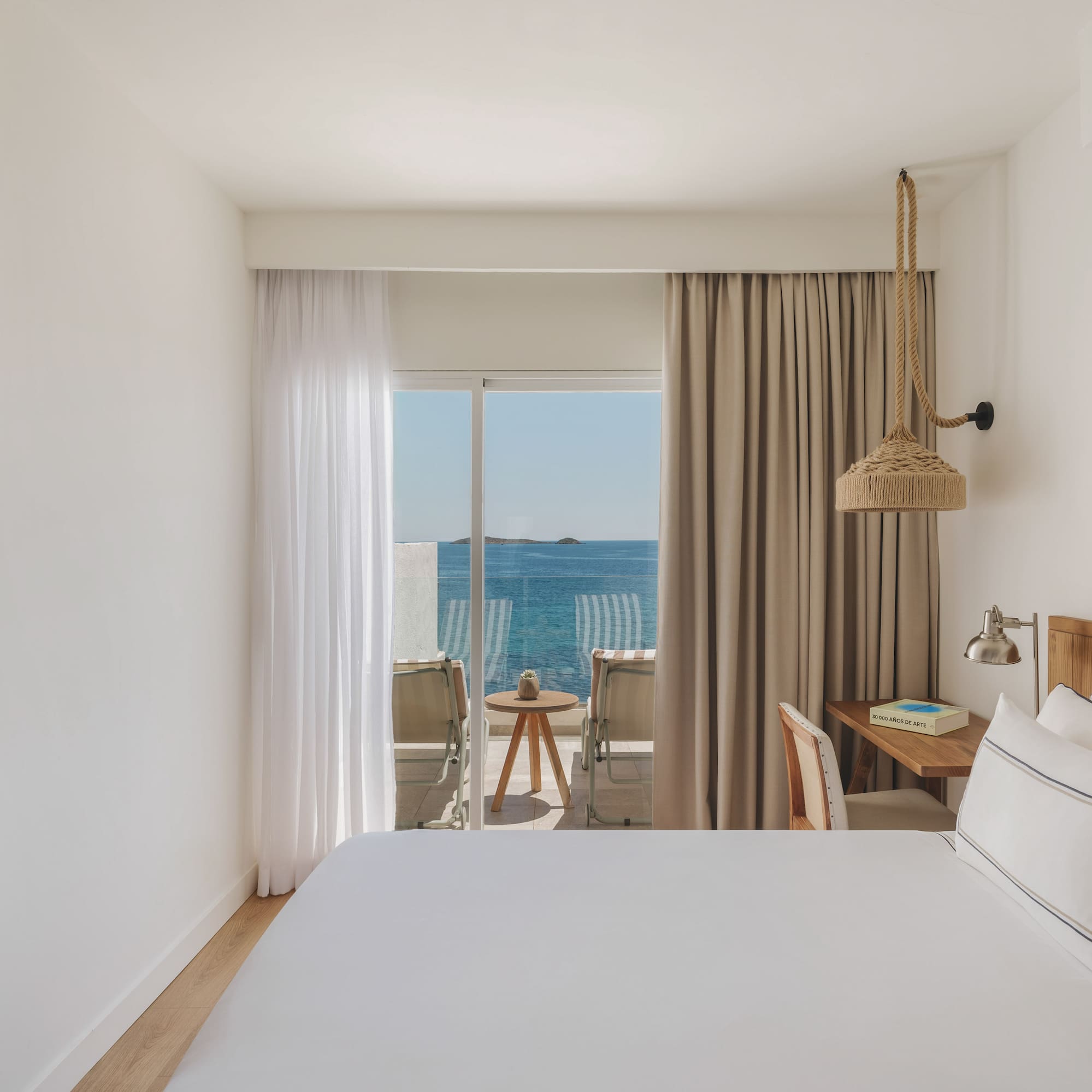 Bright hotel bedroom with sea view balcony, white bed, and wooden decor.