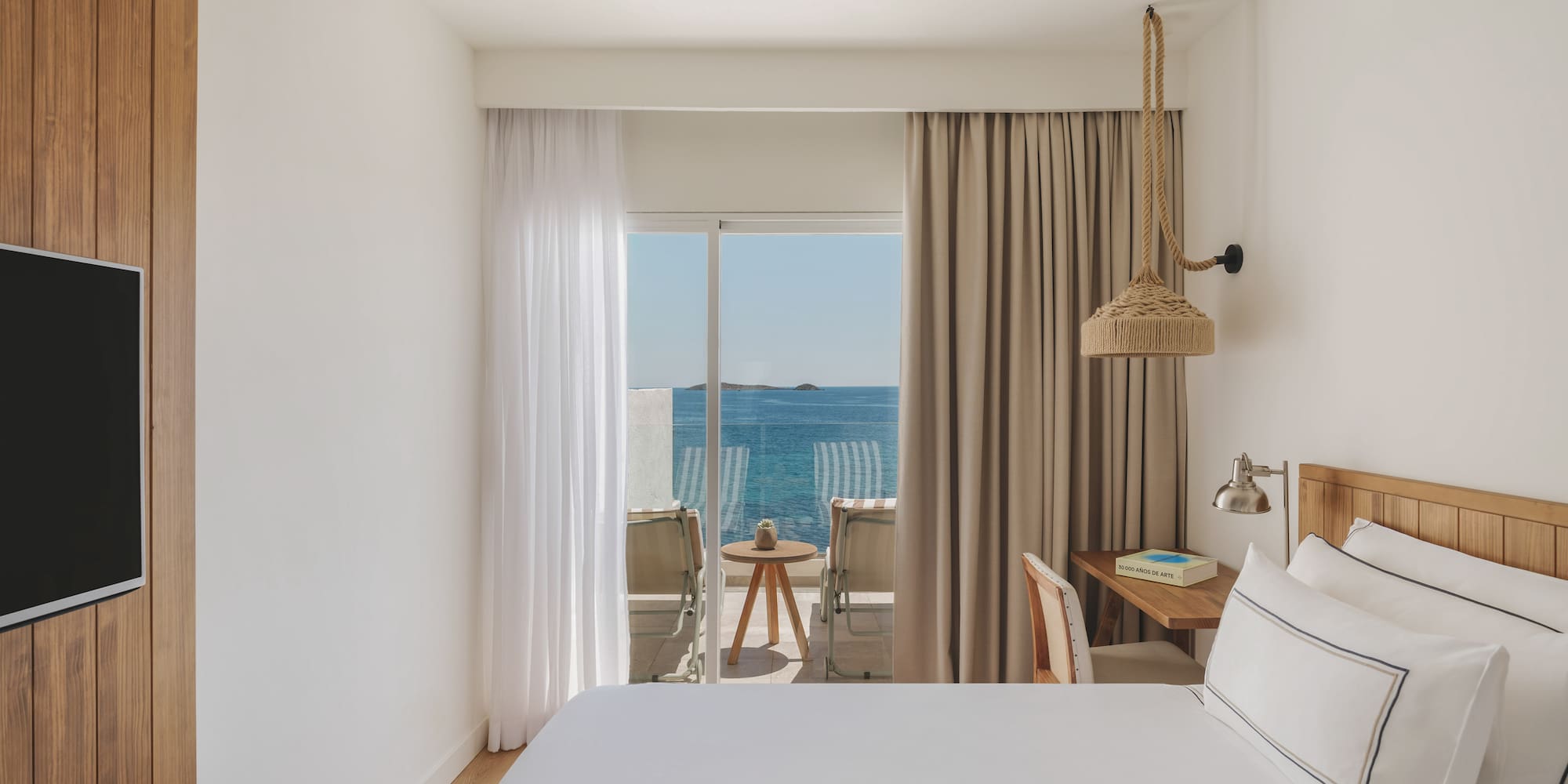 Bright hotel bedroom with sea view balcony, white bed, and wooden decor.