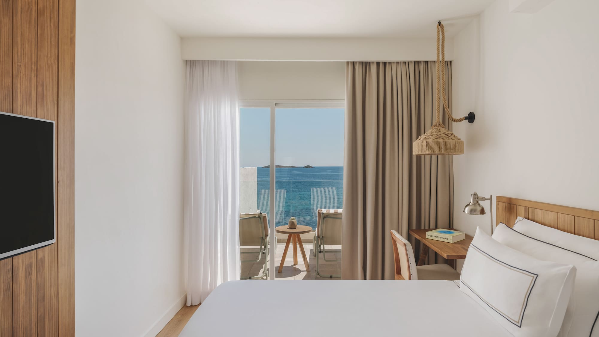 Bright hotel bedroom with sea view balcony, white bed, and wooden decor.